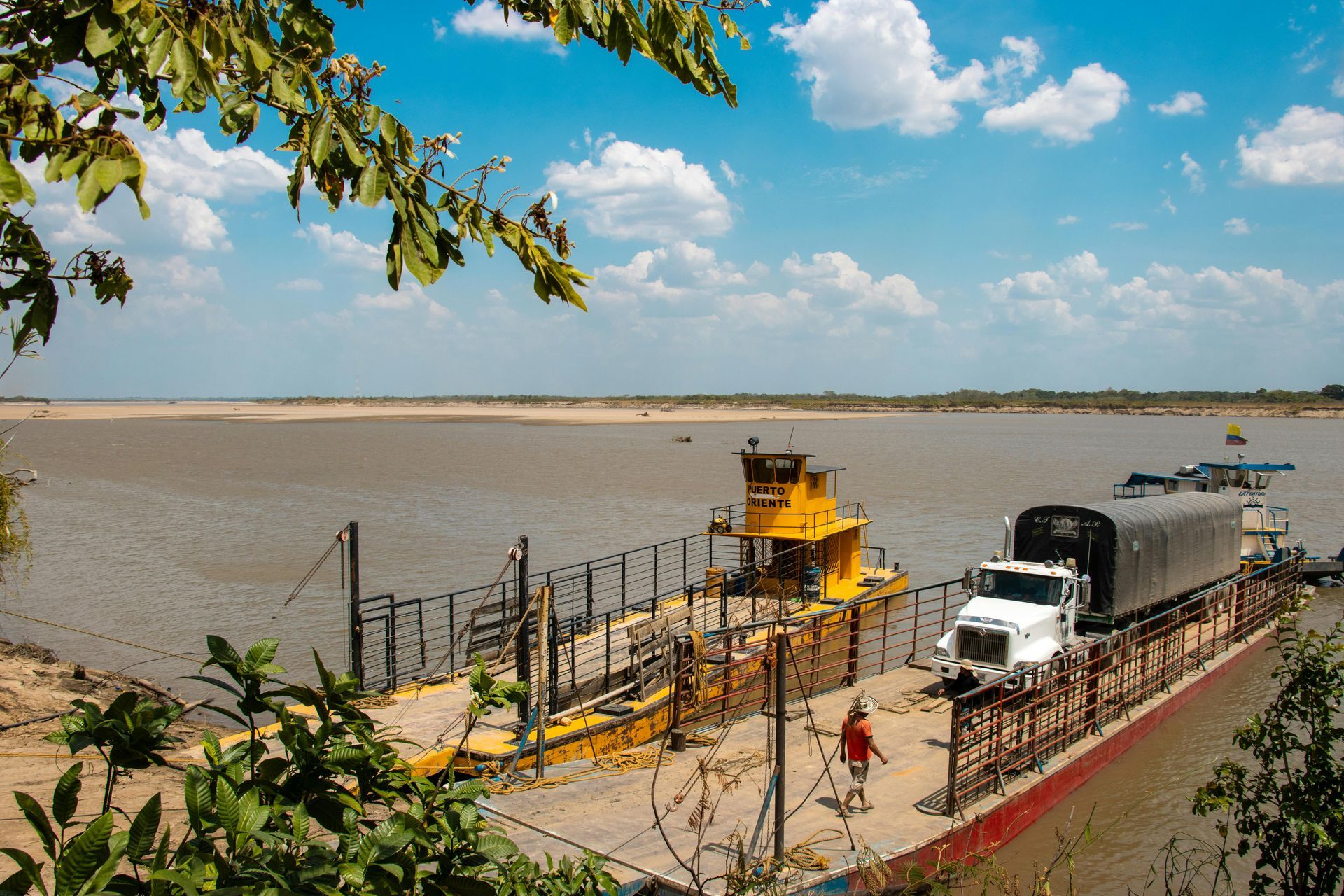 Ferry with yellow barge transports a white truck across a wide river under a blue sky.