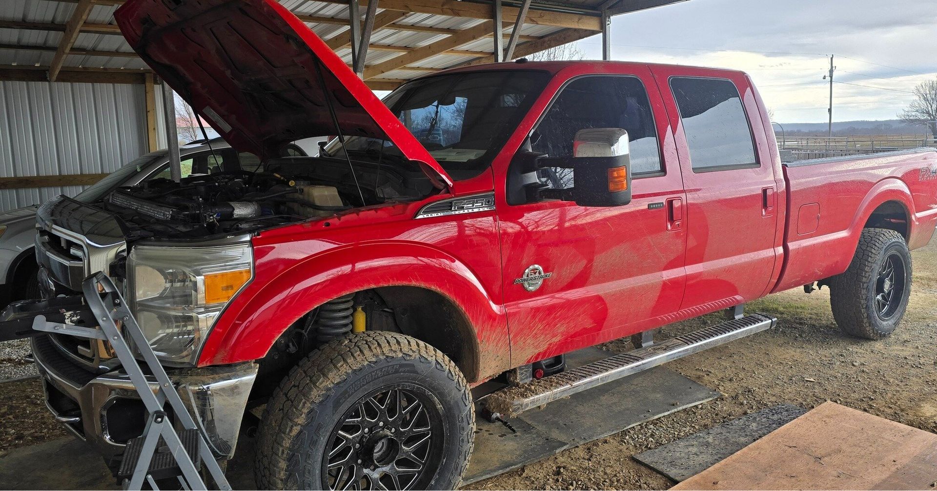 A red truck with the hood up is parked in a dirt lot.