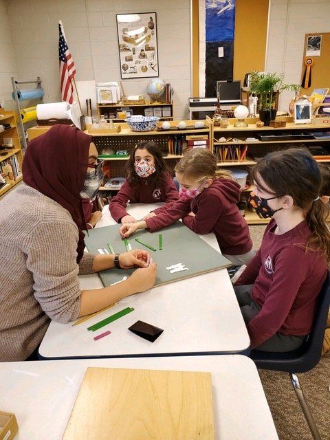 Montessori guide with three masked students working on a craft project at a table in a classroom.