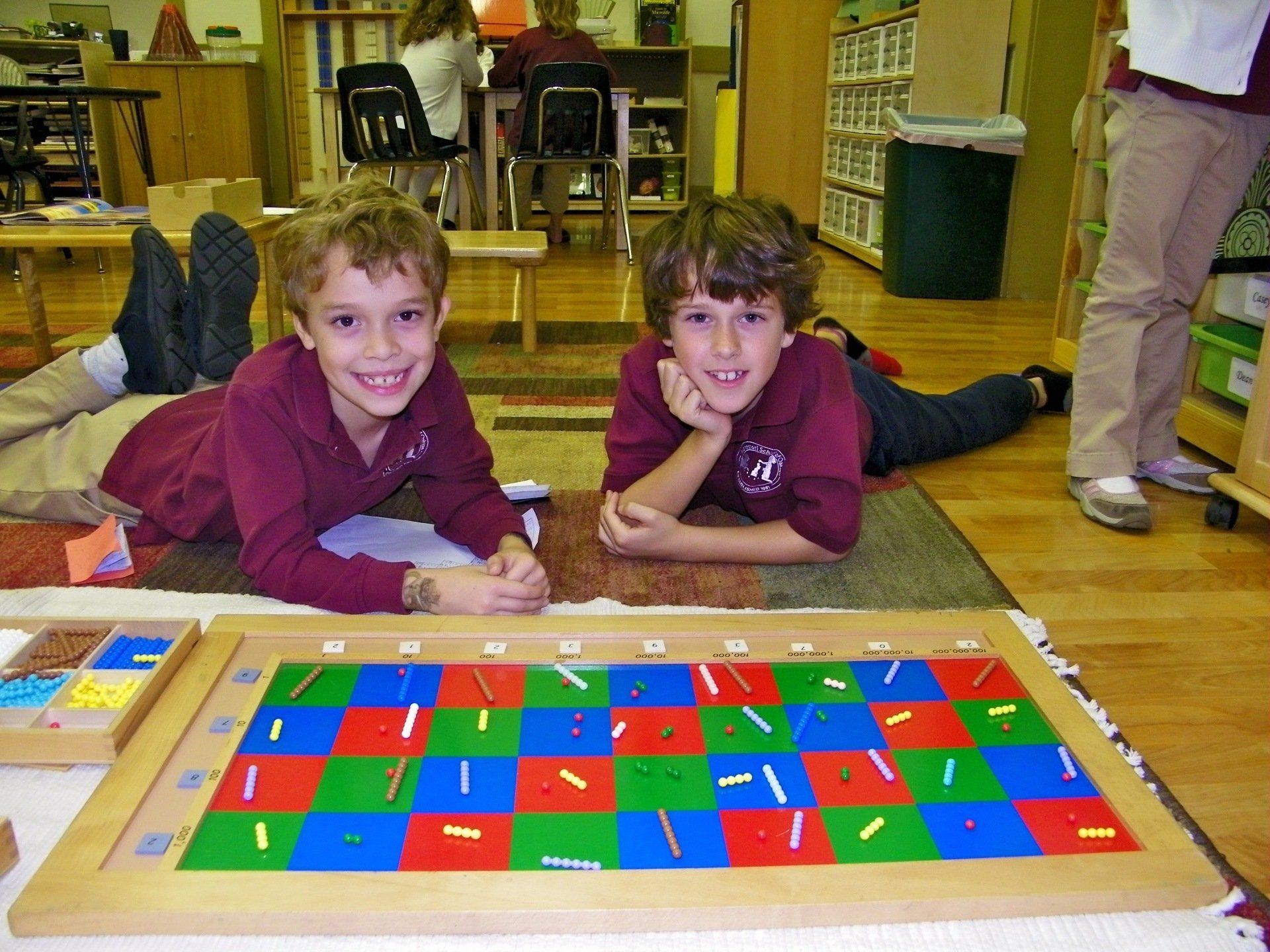 Two boys smiling to the camara while laying on the floor with Montessori Math materials in front of them