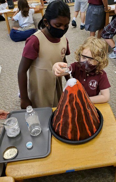 Two Montessori children in masks make a volcano erupt. One pours liquid into the volcano model.