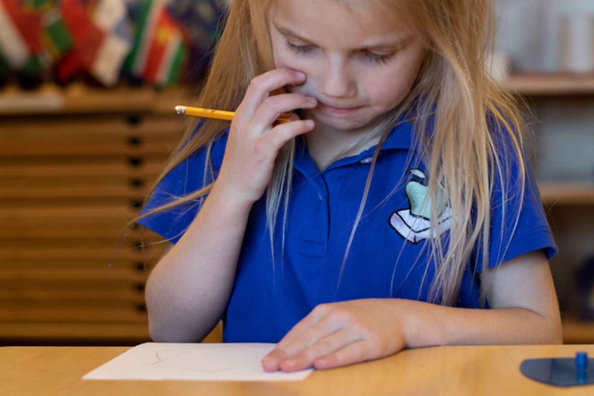 Montessori child in blue shirt, pencil to face, looking at paper on a table, focused, thoughtful expression.