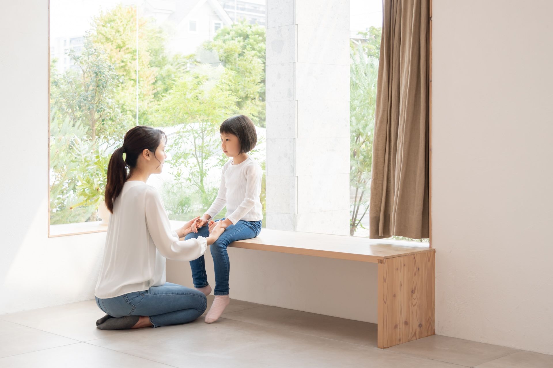 A woman is kneeling down next to a little girl sitting on a bench in front of a window.