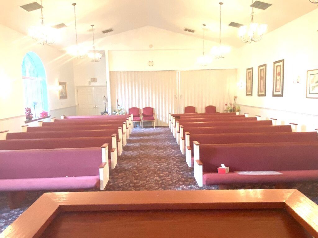 Interior of a funeral home chapel with rows of red pews, a wood altar, and framed artwork on the walls.