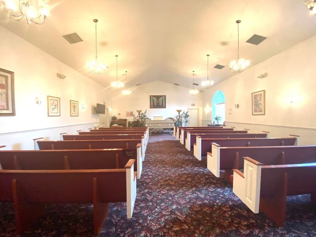 Interior of a chapel with rows of pews, chandelier lighting, and a floral arrangement on a central table.