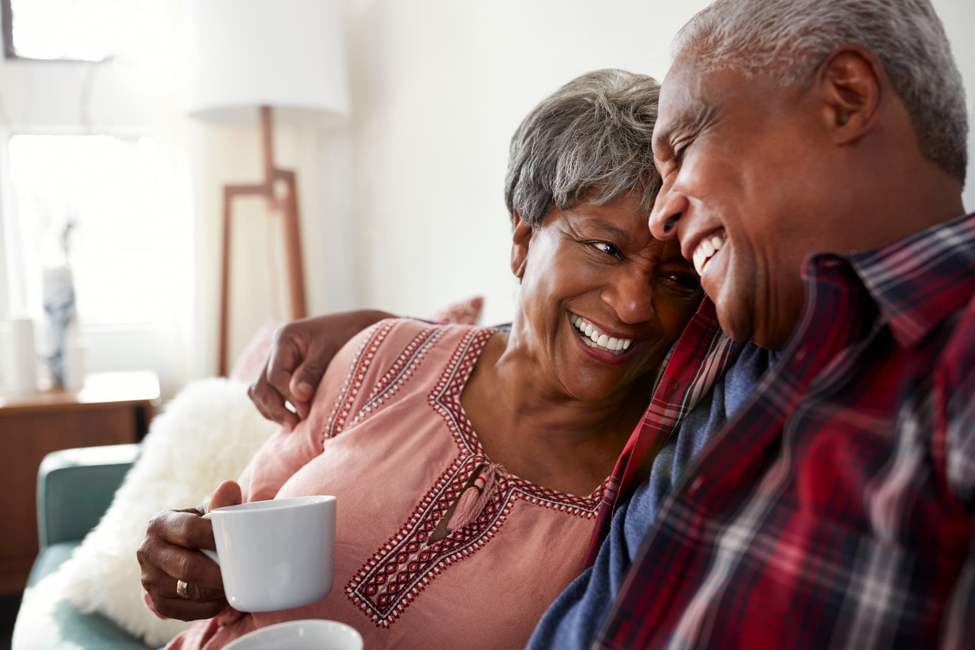 Couple smiling, embracing on a sofa. One holds a white mug, enjoying a relaxed moment at home.