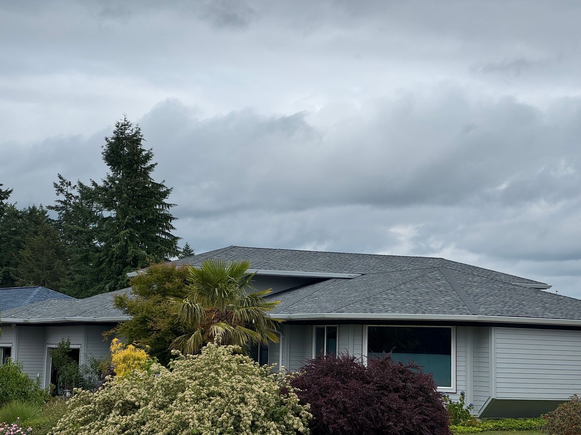 A house with a gray roof is surrounded by bushes and trees on a cloudy day.