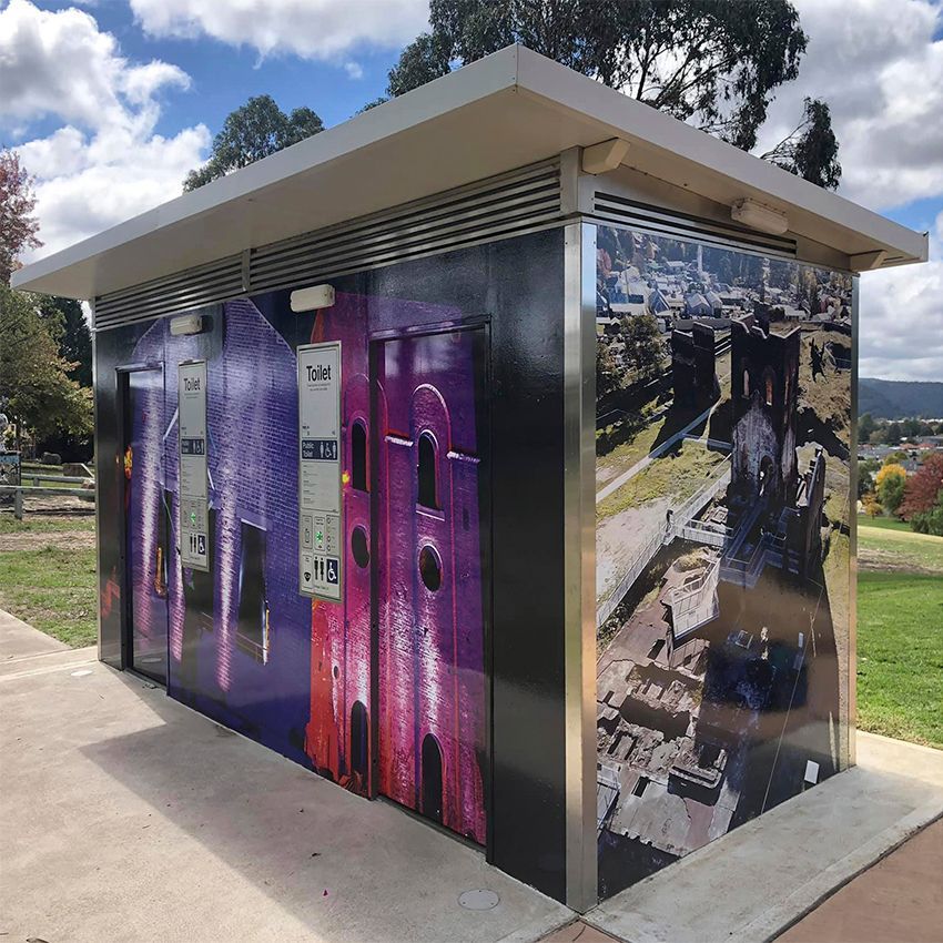 Public toilet with exterior artwork in a park setting. The toilet has a neutral roof, stainless steel corners, and walls