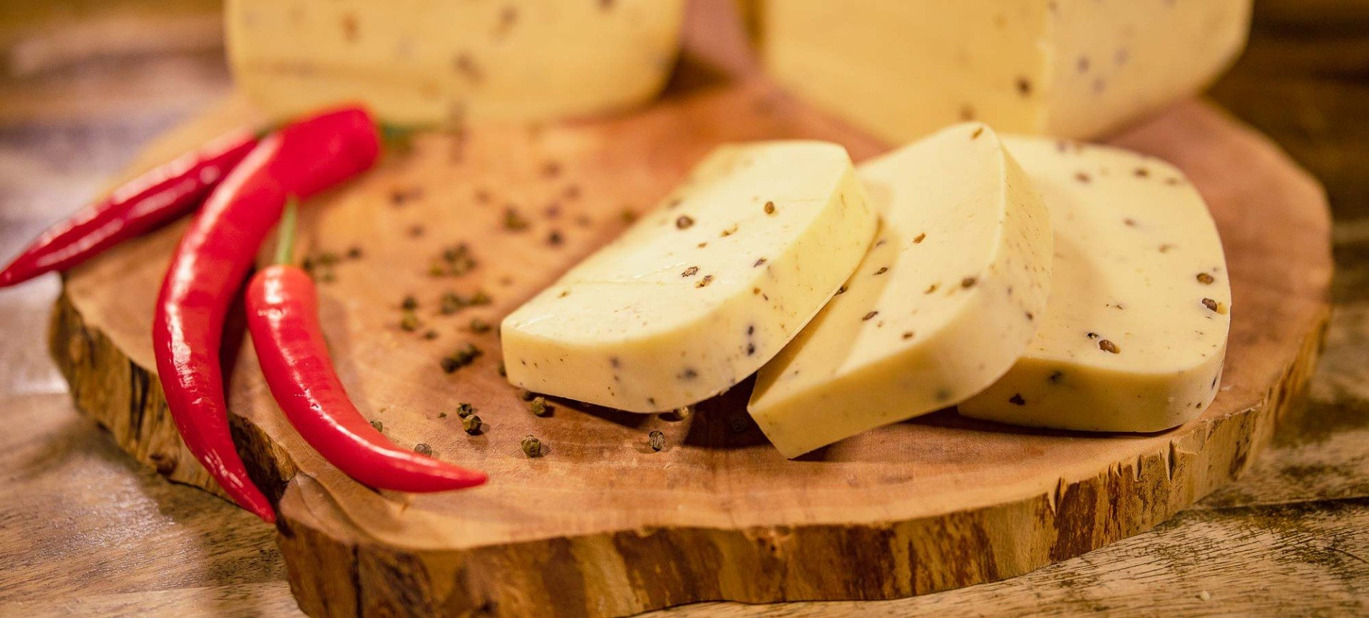 Nahaufnahme: Ein hölzernes Schneidebrett mit angeschnittenem Hofkäse mit Pfeffer. Daneben Pfefferkörner und rote Peperoni.
