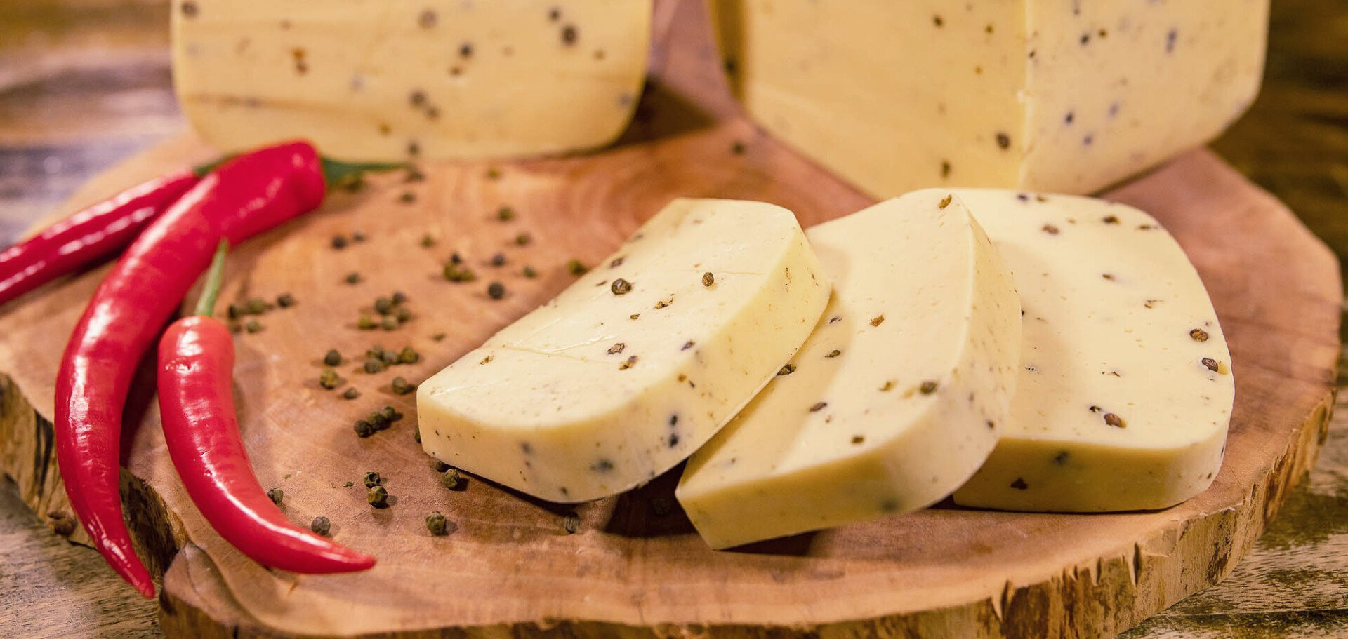 Nahaufnahme: Ein hölzernes Schneidebrett mit angeschnittenem Hofkäse mit Pfeffer. Daneben Pfefferkörner und rote Peperoni.