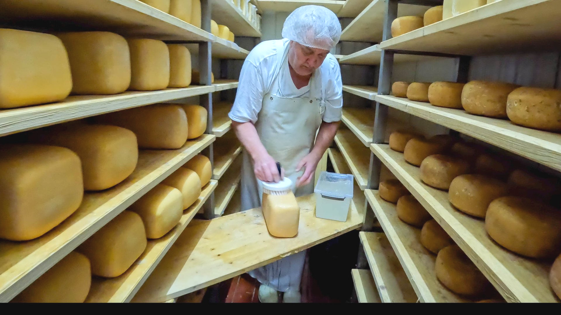 Ein Mann in einer weißen Schürze steht in einem Raum voller Käseregale und bestreicht einen Laib Käse auf einem Holzbrett.