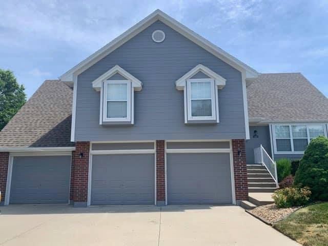 Two-story house with gray siding, three-car garage, and brown roof under a partly cloudy sky.