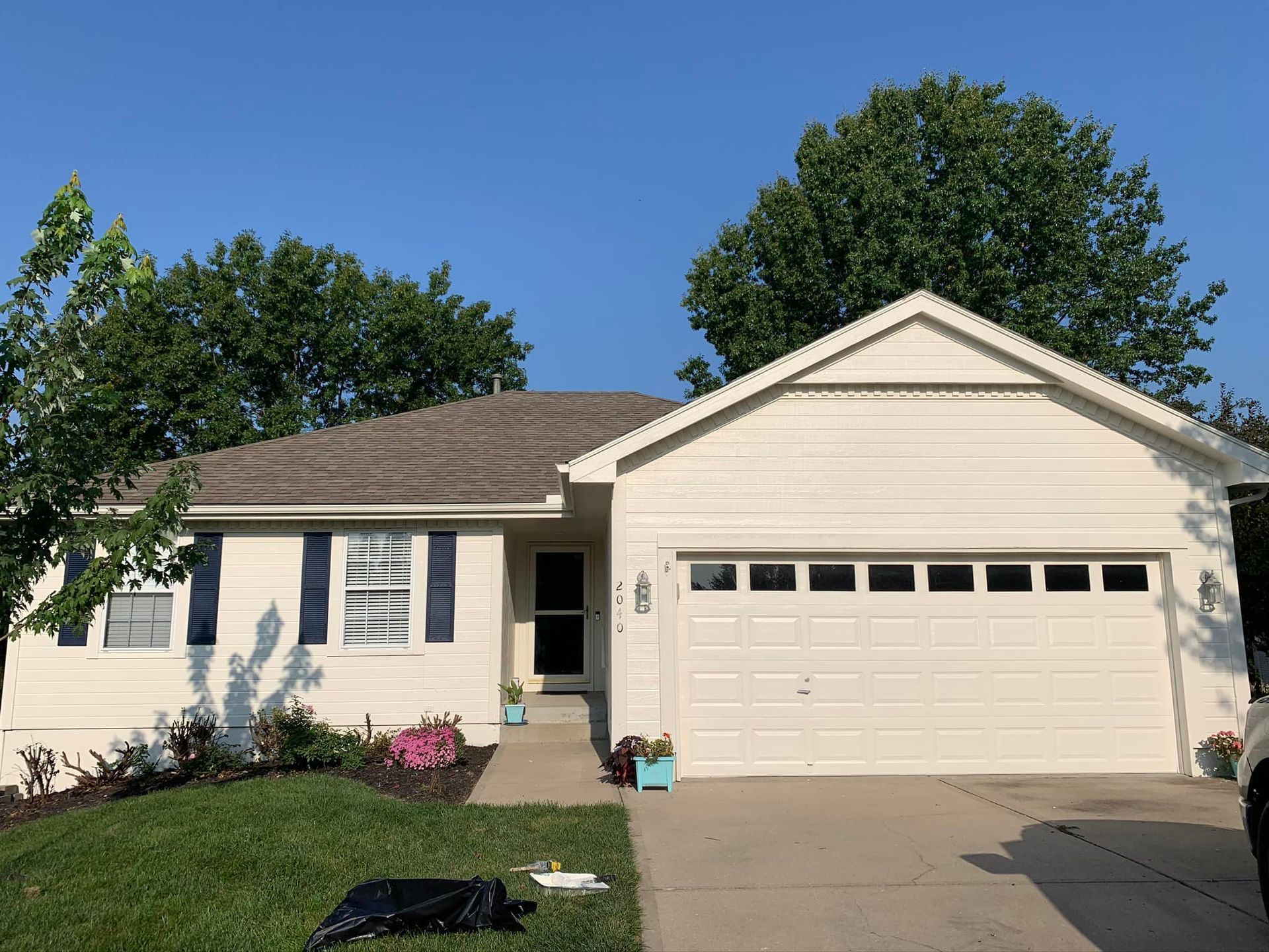 Cream-colored house with a garage, dark blue shutters, brown roof, and green lawn under a blue sky.