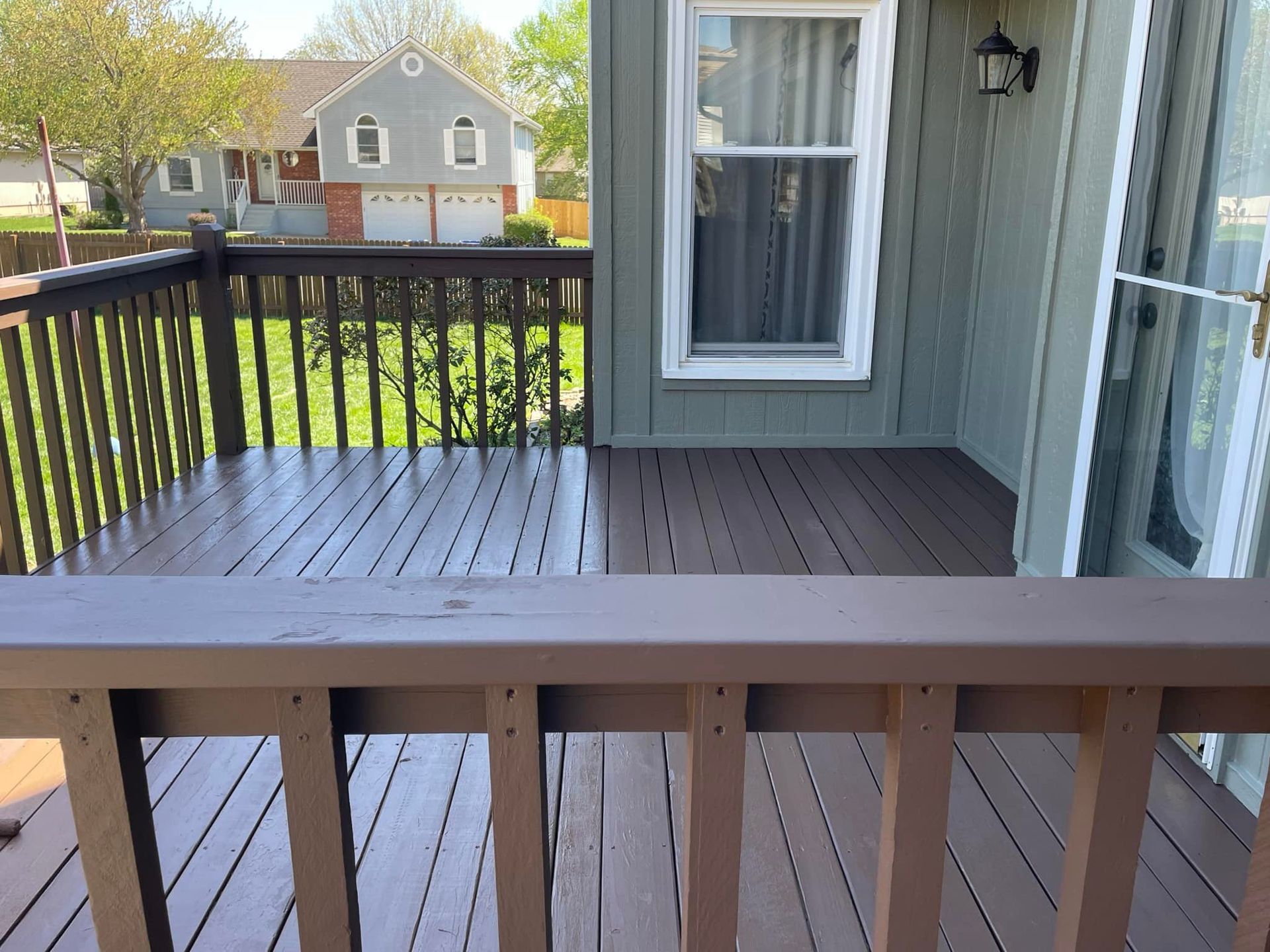 Brown painted deck with railing, house exterior in the background. Sunny day.