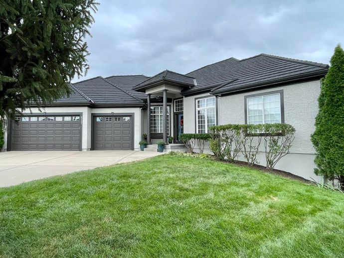 Gray stucco house with black roof, two-car garage, and green lawn under a cloudy sky.