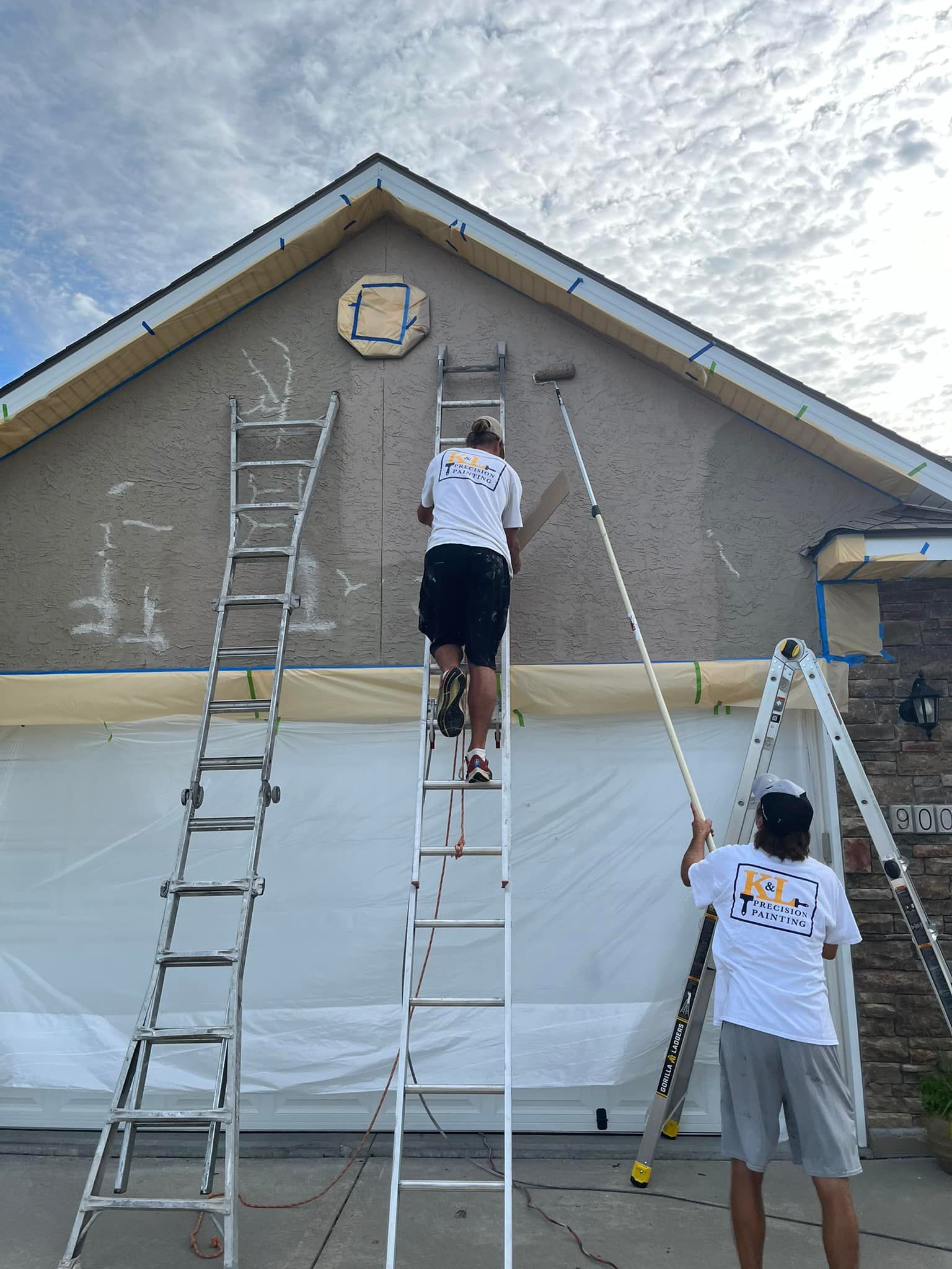Three people painting a house exterior, using ladders. Building is beige with dark roof. One person uses a long handled roller.