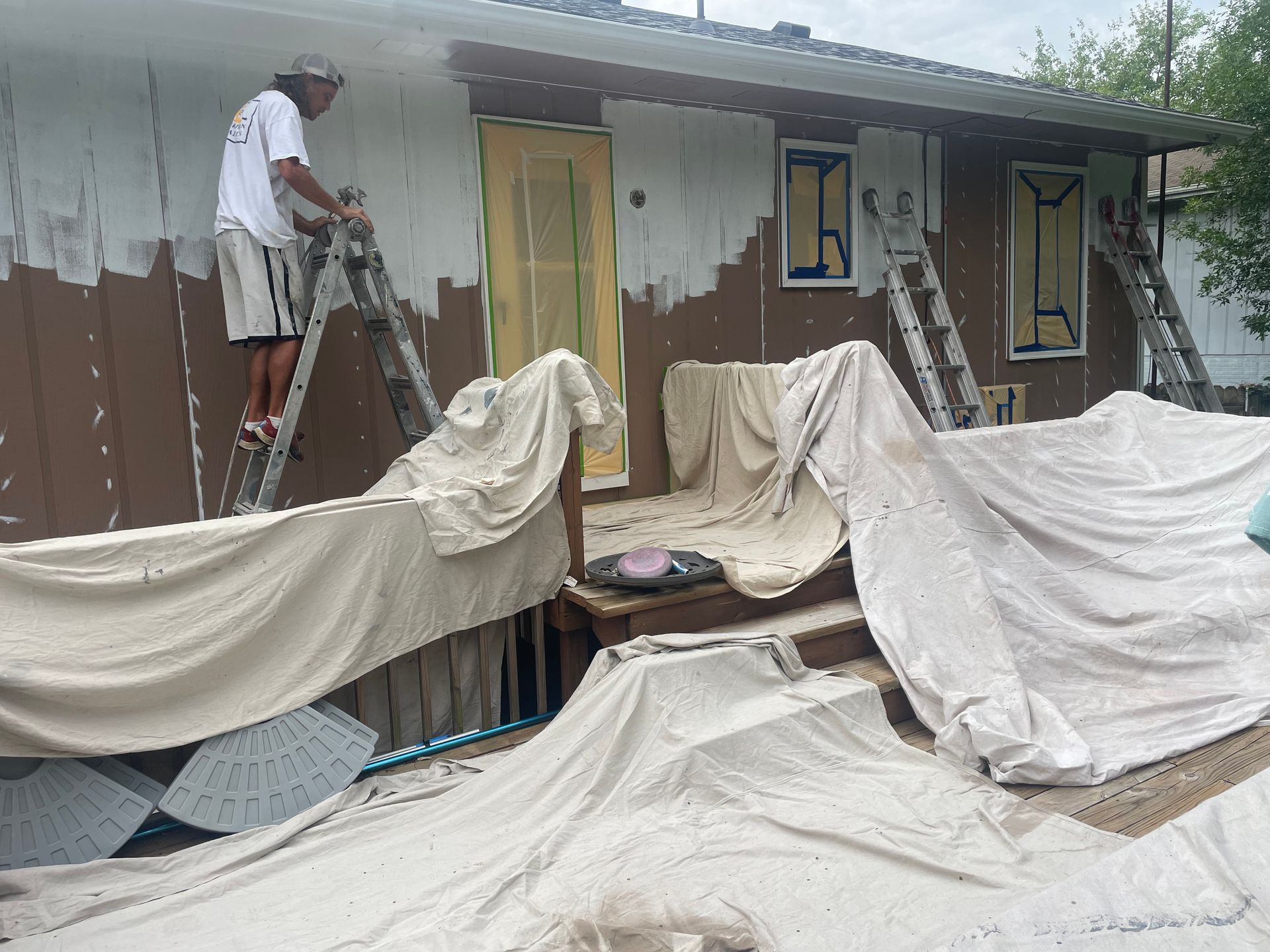 A person on a ladder paints a brown house exterior white, with drop cloths covering the deck and ground.