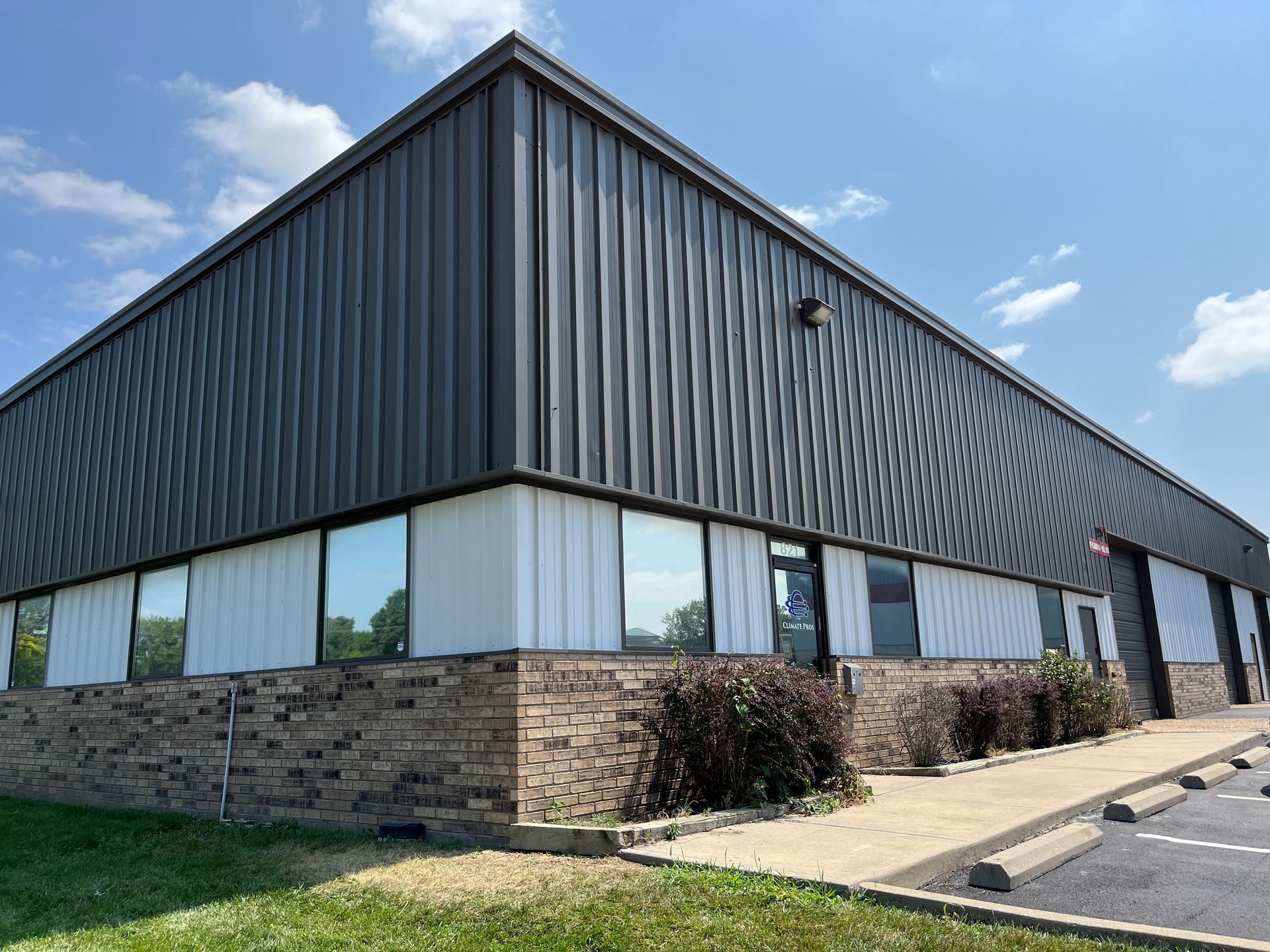 A corner view of a one-story industrial building featuring dark metal upper walls, white lower walls, and a brick base.