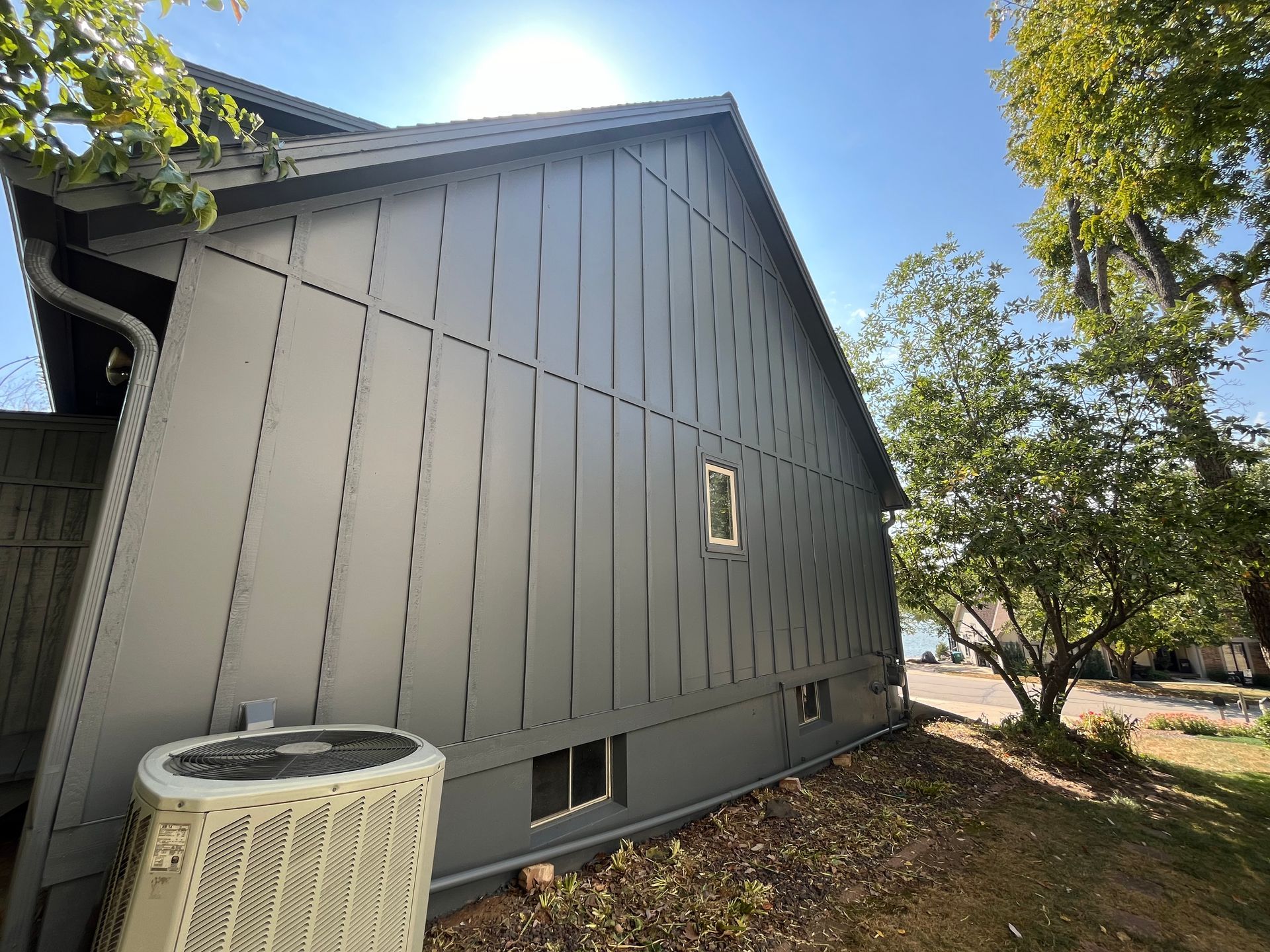 Side view of a house with grey board-and-batten siding, a small window, and an outdoor air conditioning unit.