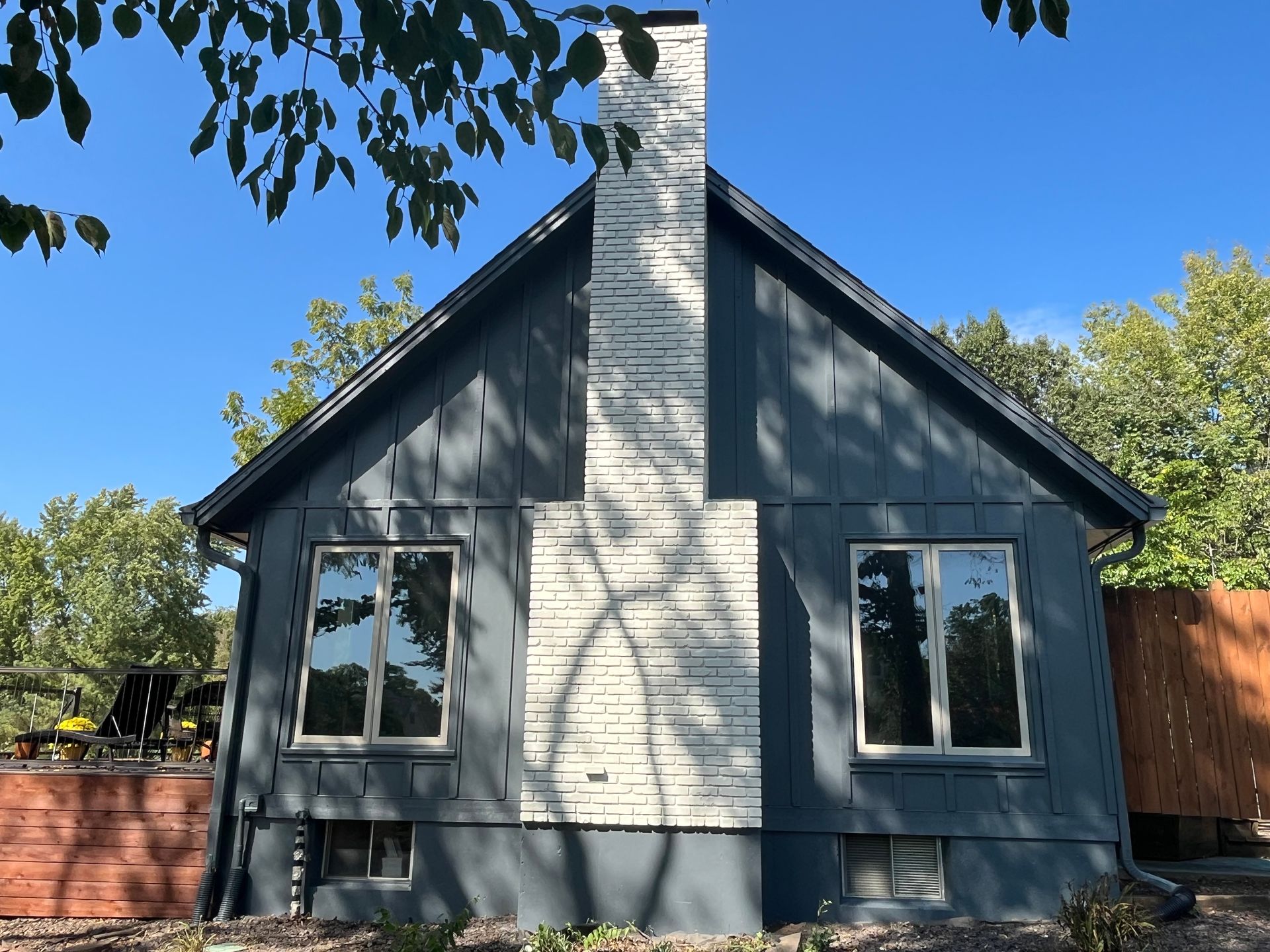 A grey house with a white stone chimney and two windows, set against a bright blue sky with surrounding trees.