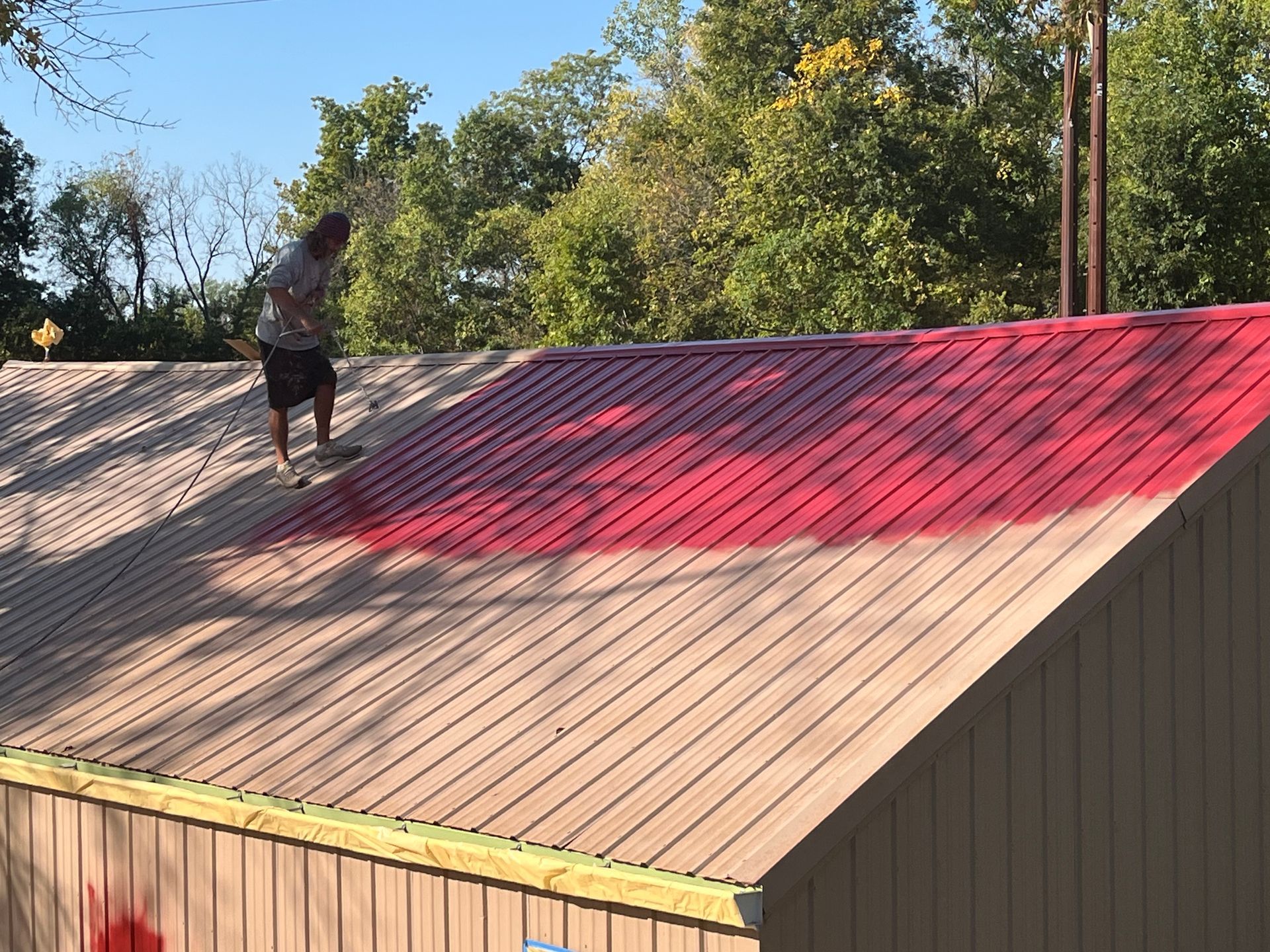 A person walks on a slanted metal roof, painting a section of it bright red while the rest remains a light tan.