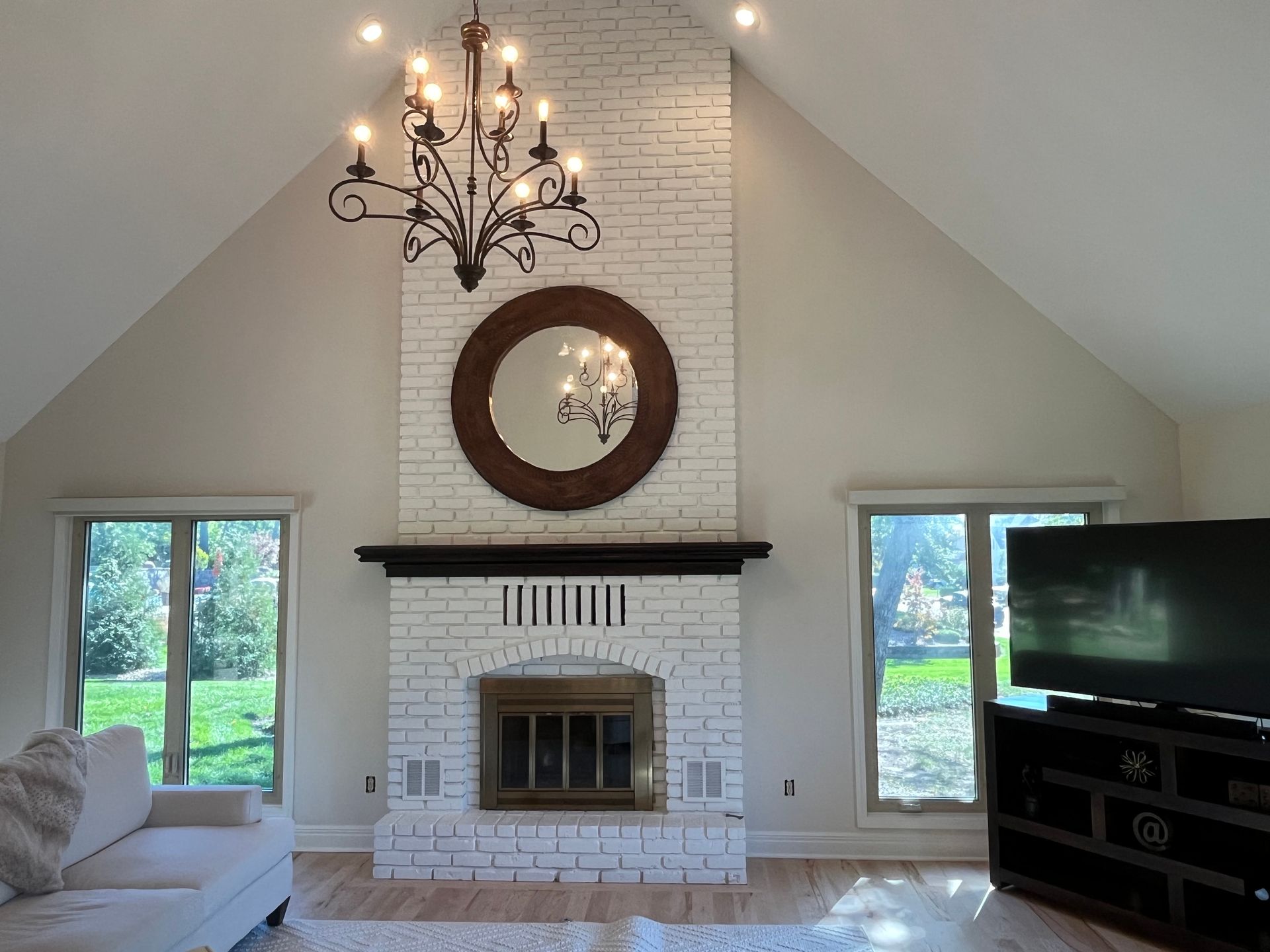 A white brick fireplace with a dark mantel and circular mirror, centered in a room with a vaulted ceiling and chandelier.