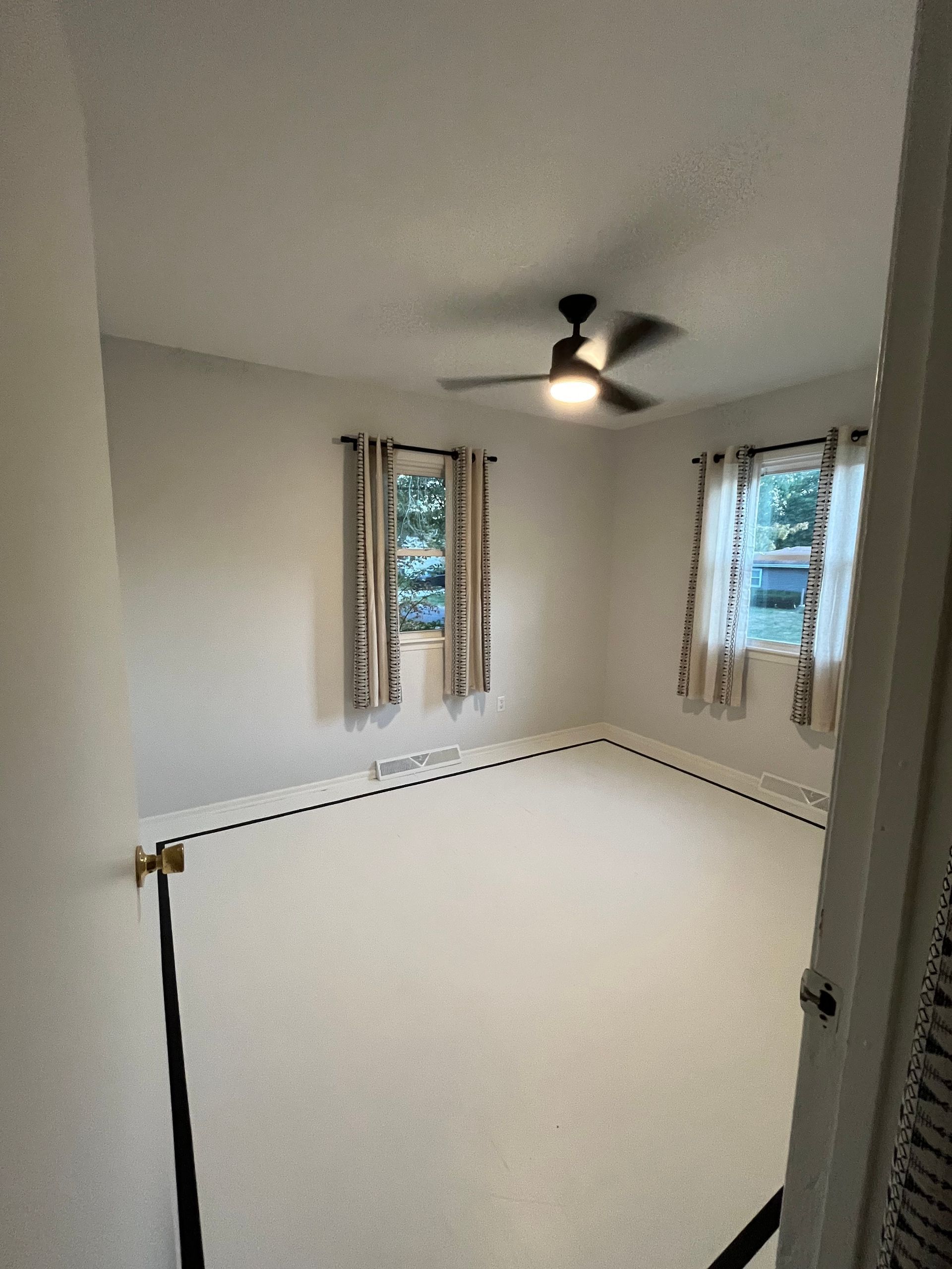 An empty bedroom with light-colored carpet, white walls, a ceiling fan, and two windows with patterned curtains.