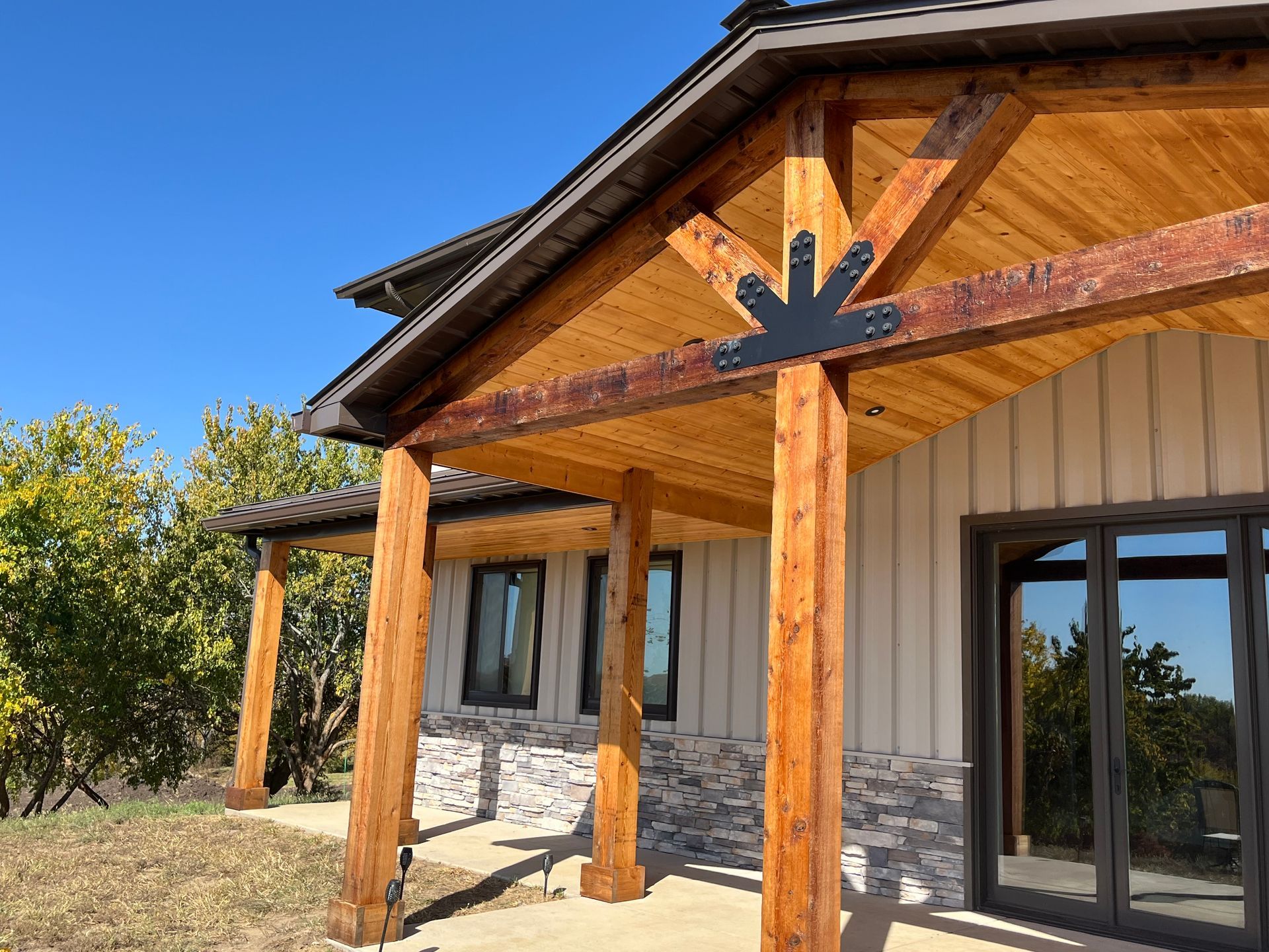 Covered porch with timber posts, exposed wood ceiling, black sunburst bracket, and stone siding under a clear blue sky.