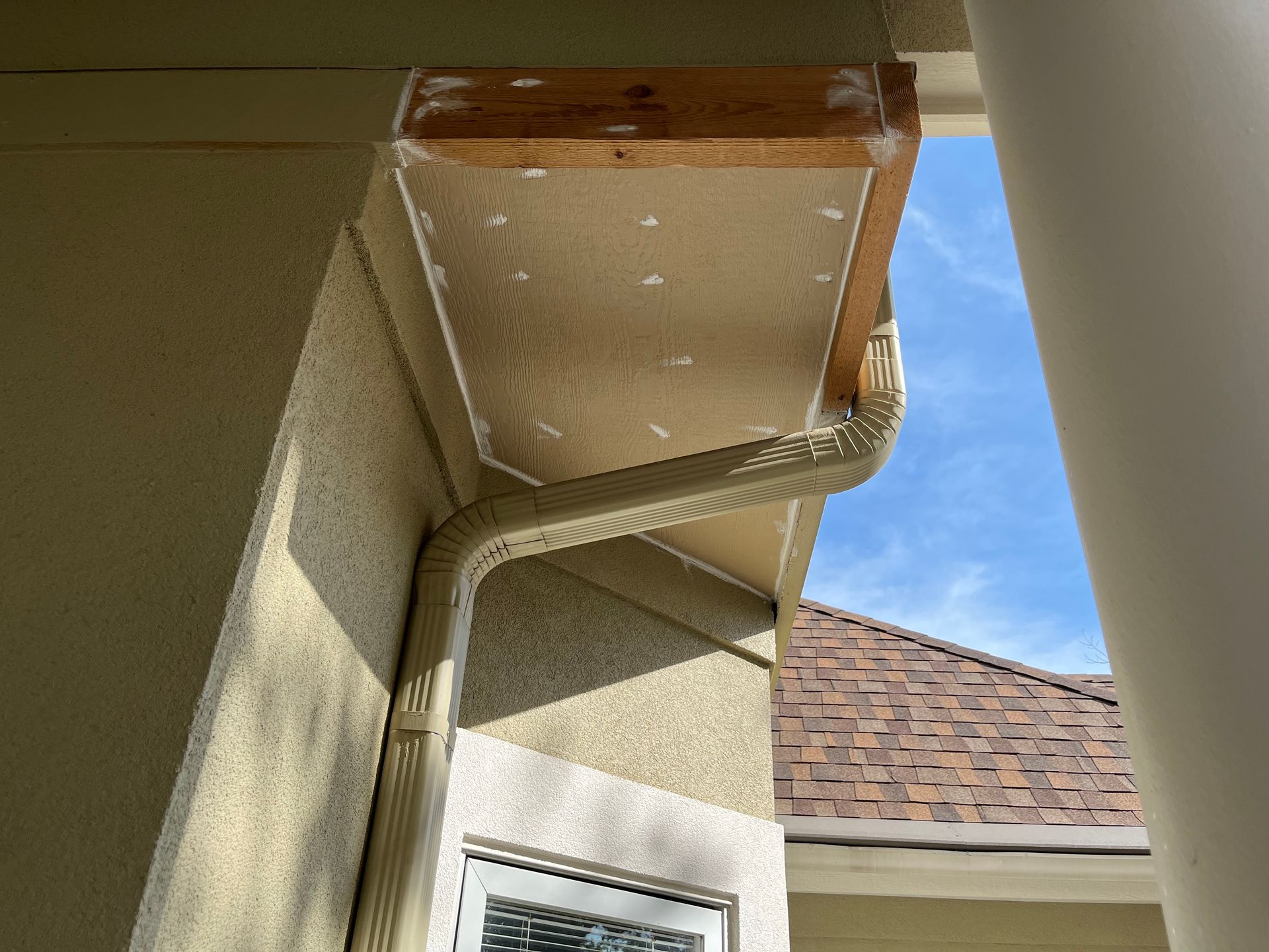 Exterior view of a beige downspout attached to a home, leading up to an unfinished, exposed wooden soffit area.