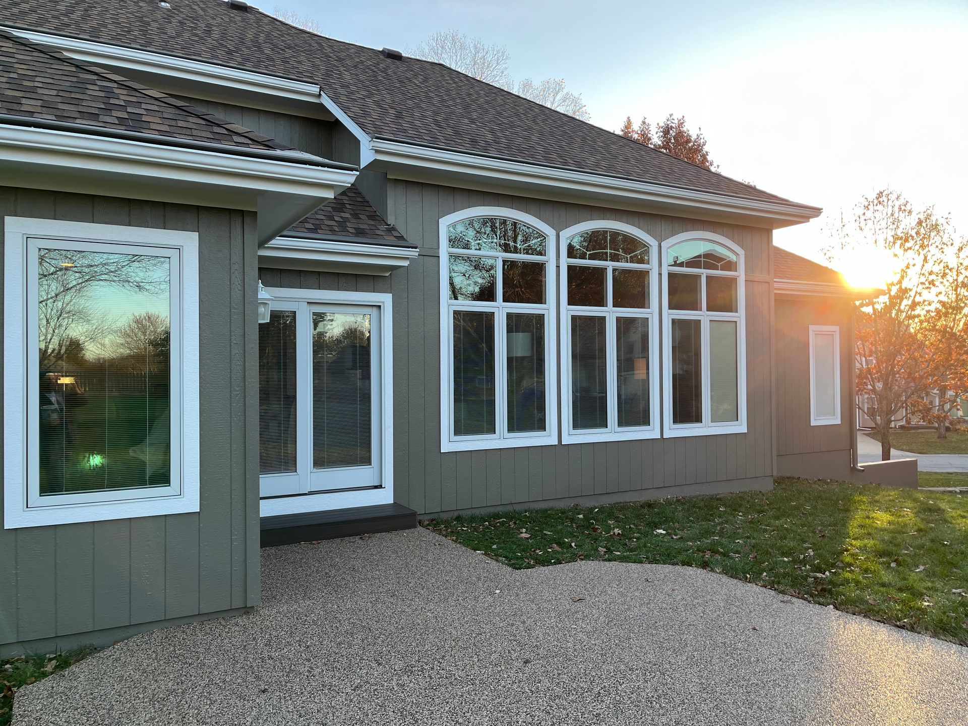 The exterior of a gray house with white-framed windows, a glass door, and a gravel patio at sunset.