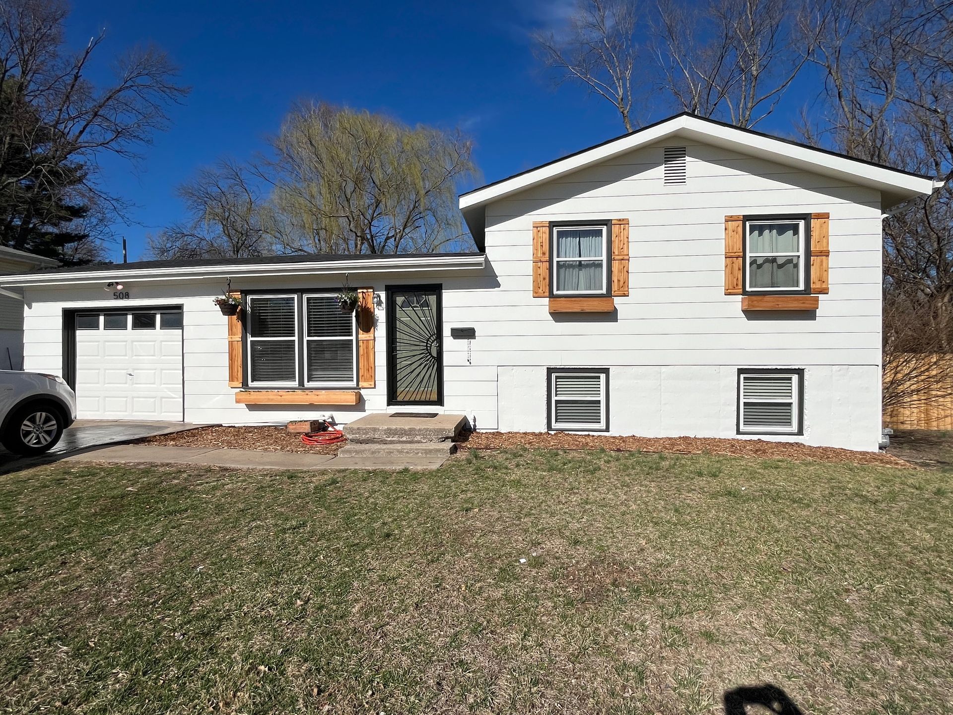 A white, split-level house with brown shutters, a single-car garage, and a lawn under a clear blue sky.