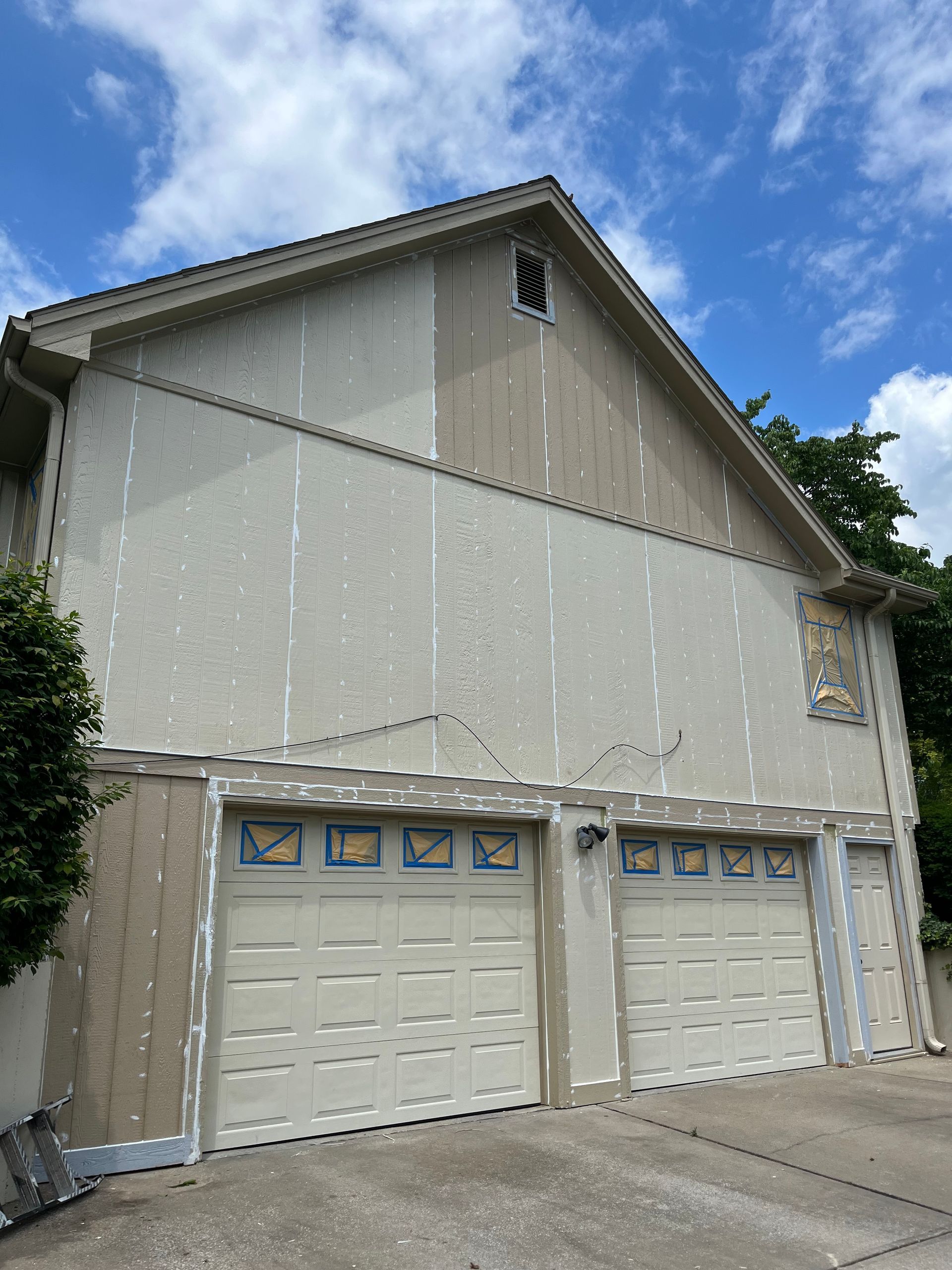 A two-story garage with light-colored siding currently undergoing a painting project with tape visible on the edges.