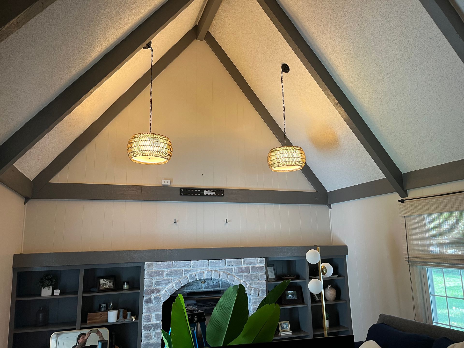 A vaulted ceiling with exposed gray beams, two hanging pendant lights, a brick fireplace, and built-in gray bookshelves.