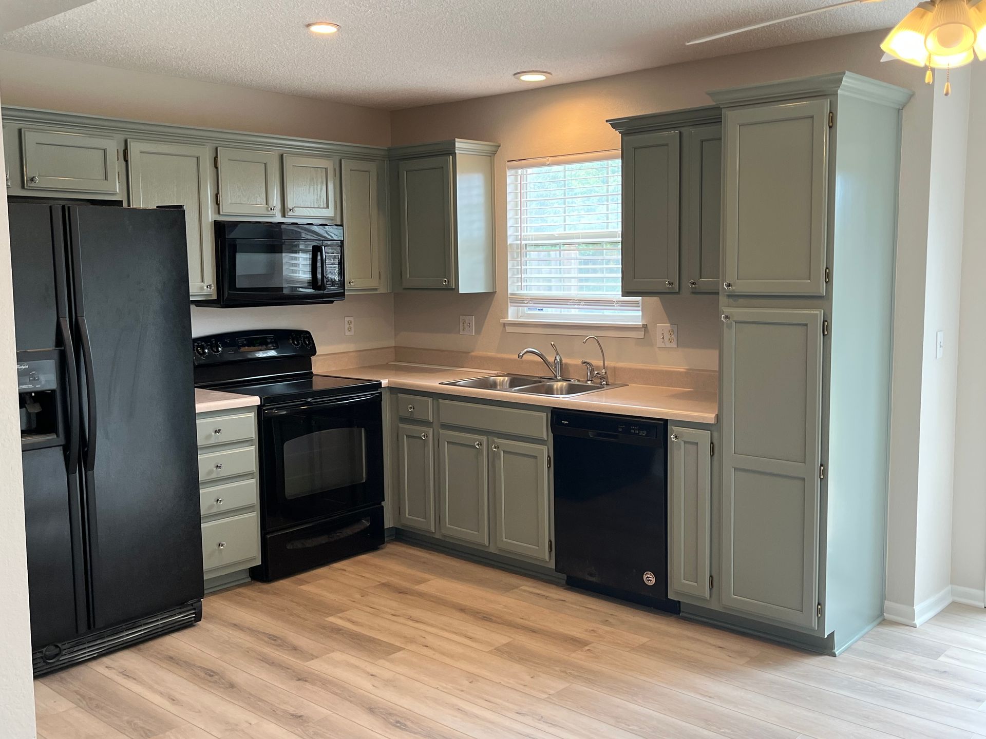 A kitchen with sage green cabinets, black appliances, light wood-style flooring, and a window above the sink.