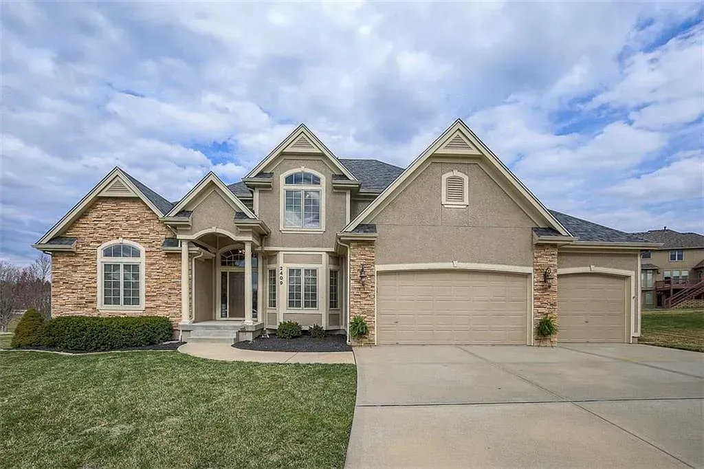 A two-story suburban house with stone and beige siding, a three-car garage, and a large front lawn under a cloudy sky.