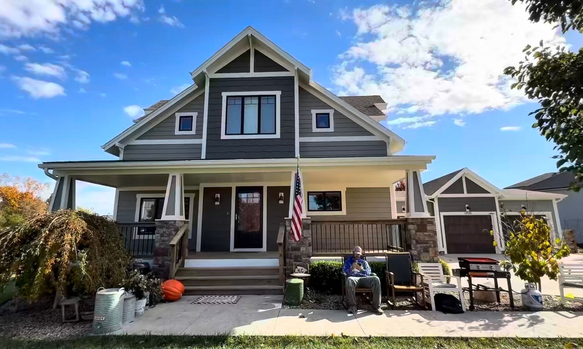 A person sitting on the front porch of a two-story grey house with a wrap-around porch and an attached garage.