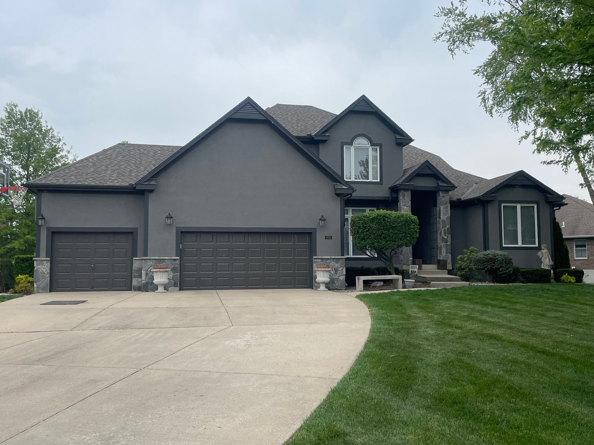 A two-story, gray house with a three-car garage, stone accents, and a large front lawn under a cloudy sky.