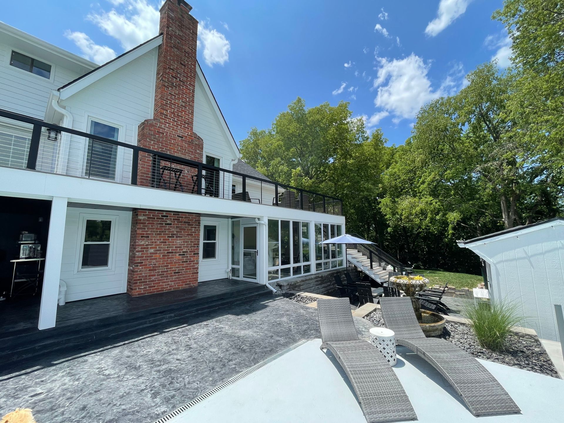 A house exterior with a white deck, brick chimney, gray paved patio, and two lounge chairs under a blue sky with trees.