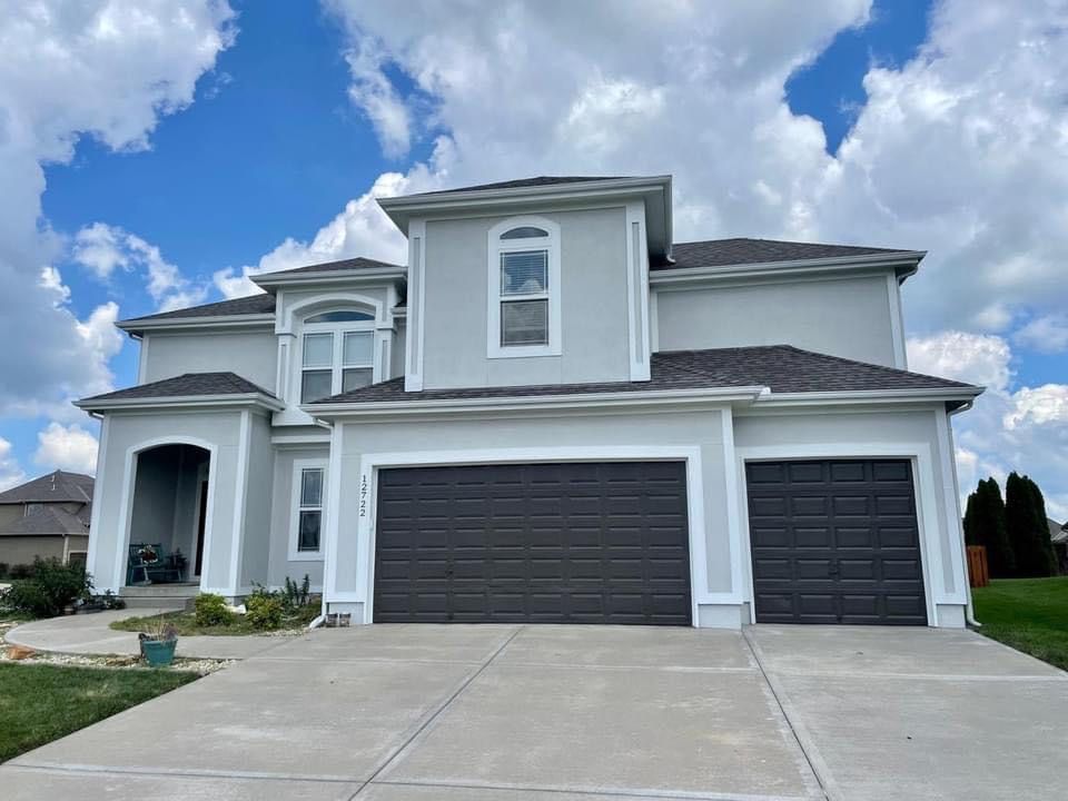A two-story light gray house with a three-car garage and a paved driveway under a blue, cloudy sky.