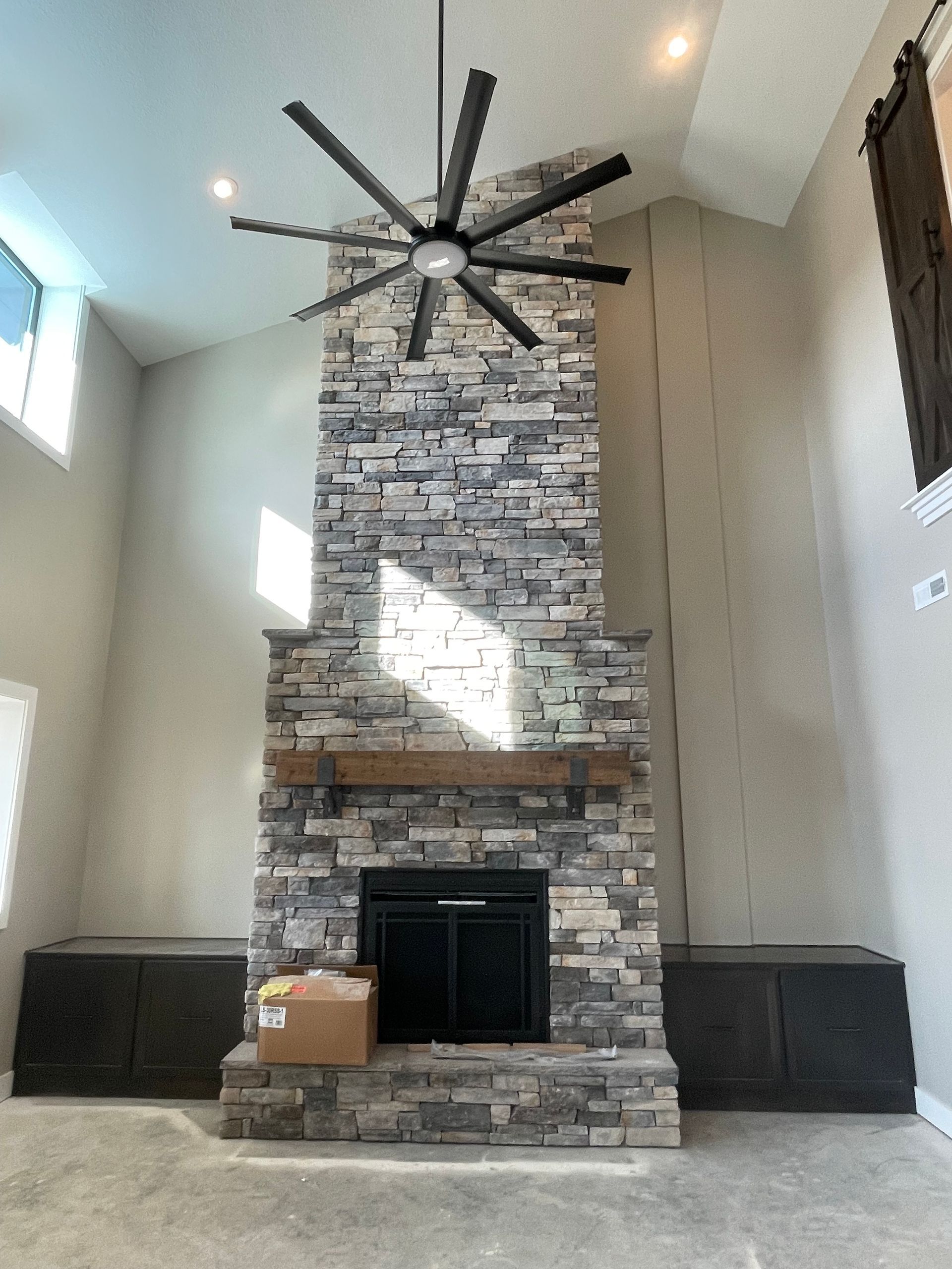 A tall stone fireplace serves as the centerpiece in a high-ceilinged room with dark cabinets and a large ceiling fan.