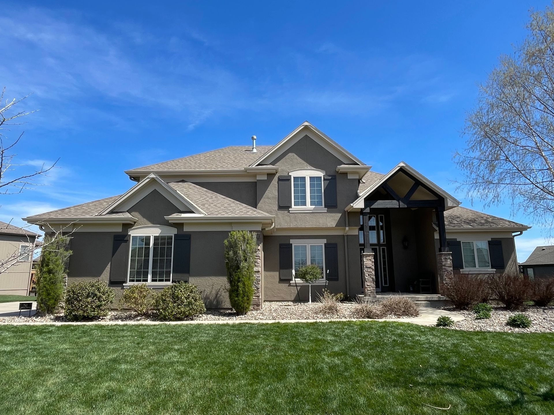 A two-story grey stucco house with dark shutters, a stone-accented entryway, and a green lawn under a bright blue sky.