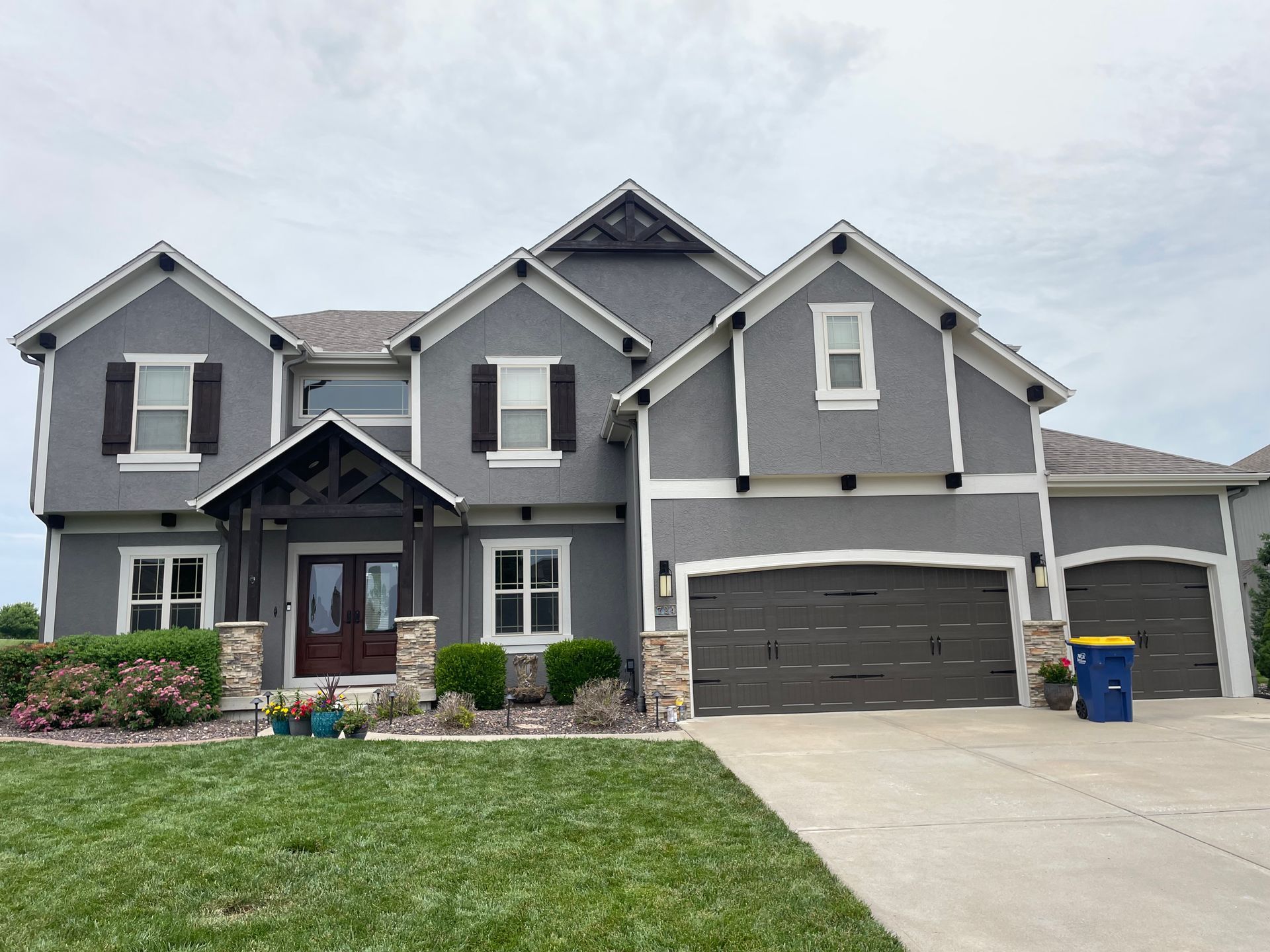 A two-story grey stucco house with a three-car garage, white trim, stone accents, and a front porch.