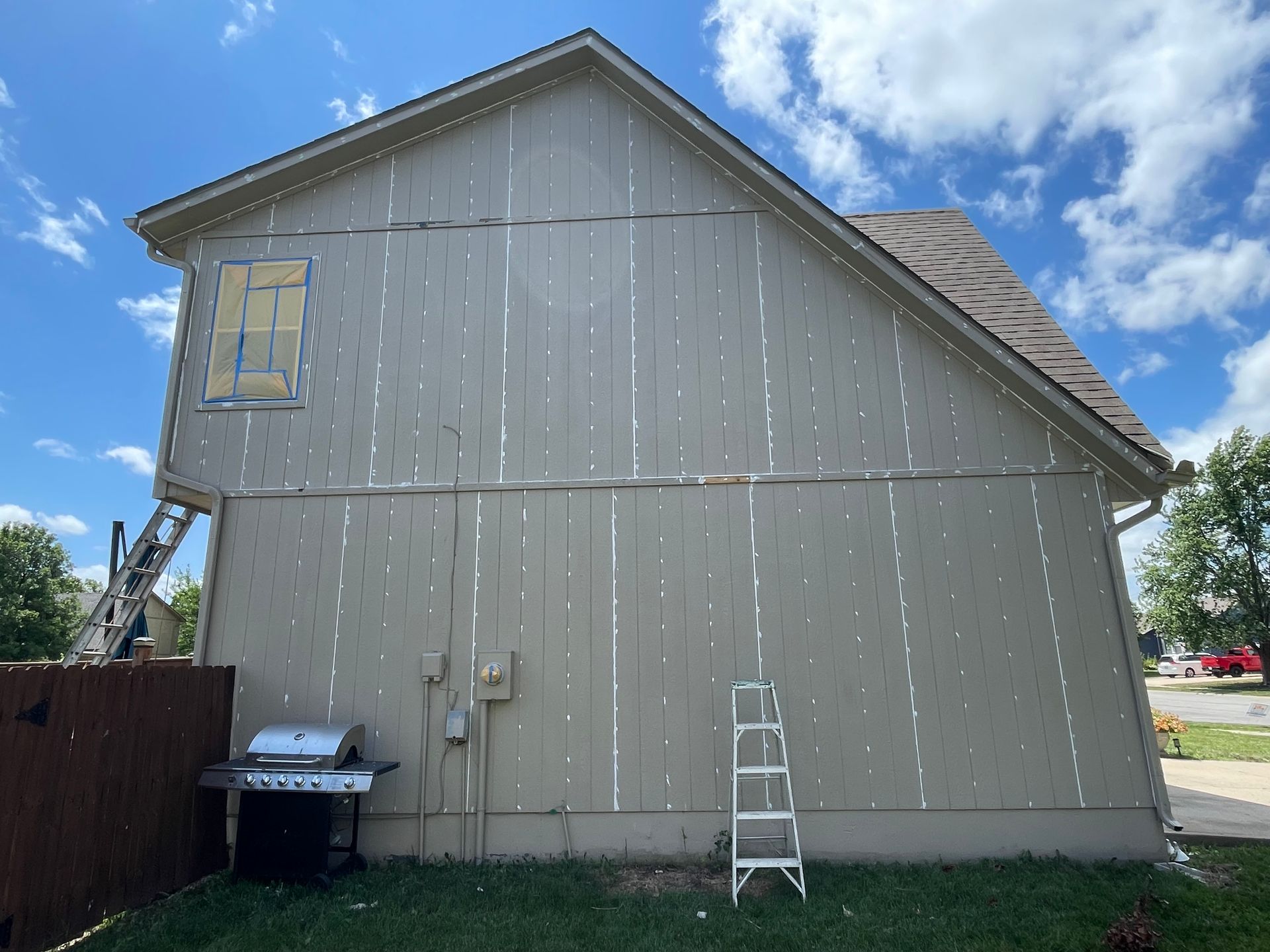Side view of a two-story house with beige vertical wood siding, a small ladder, and a grill on a sunny day.