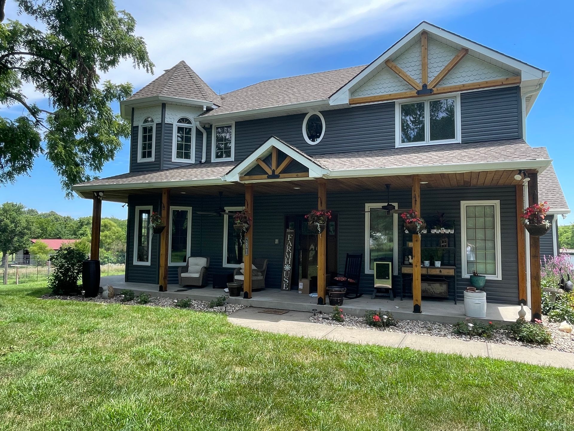 Two-story dark grey farmhouse with a wraparound porch, wooden posts, and a turret, set on a sunny, grassy lawn.