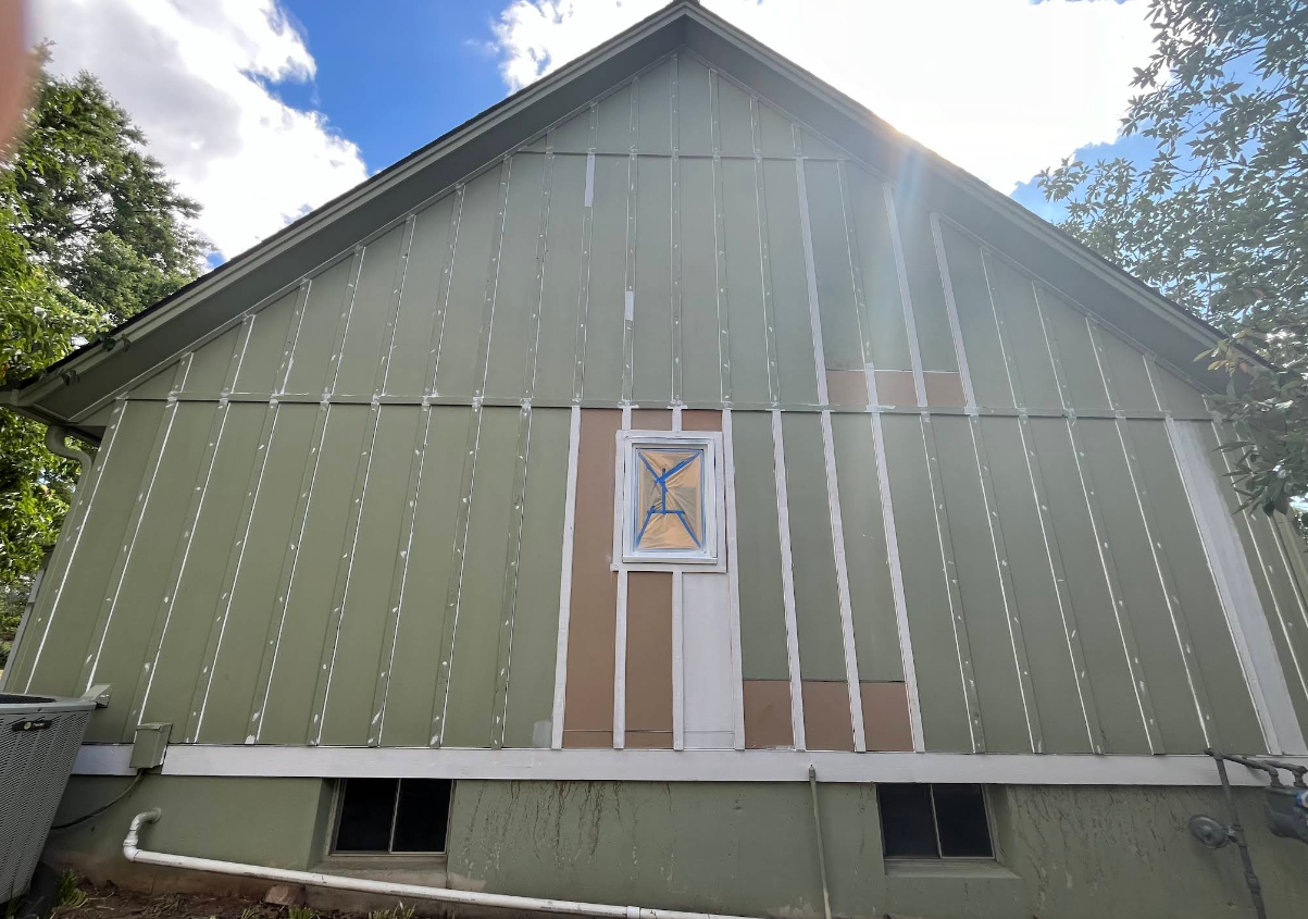 Green house siding partially removed; visible framing; blue sky.