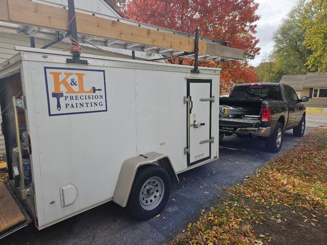 A white K&L Precision Painting utility trailer hitched to a black pickup truck, parked on a gravel driveway in autumn.