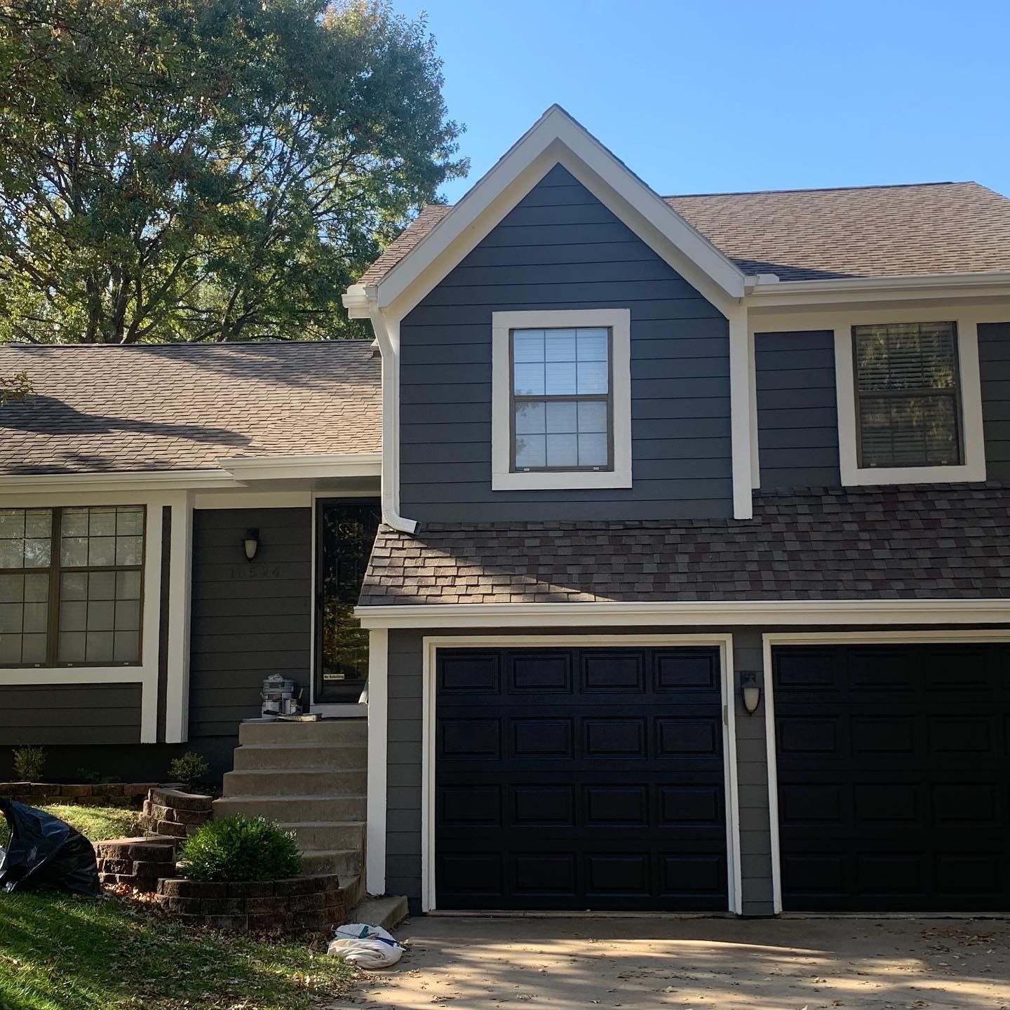 Two-story house with dark gray siding, white trim, and black garage doors. Brown roof.