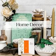 Home decor arrangement with flowers, books, candles, and a small orange door against a white wood backdrop.