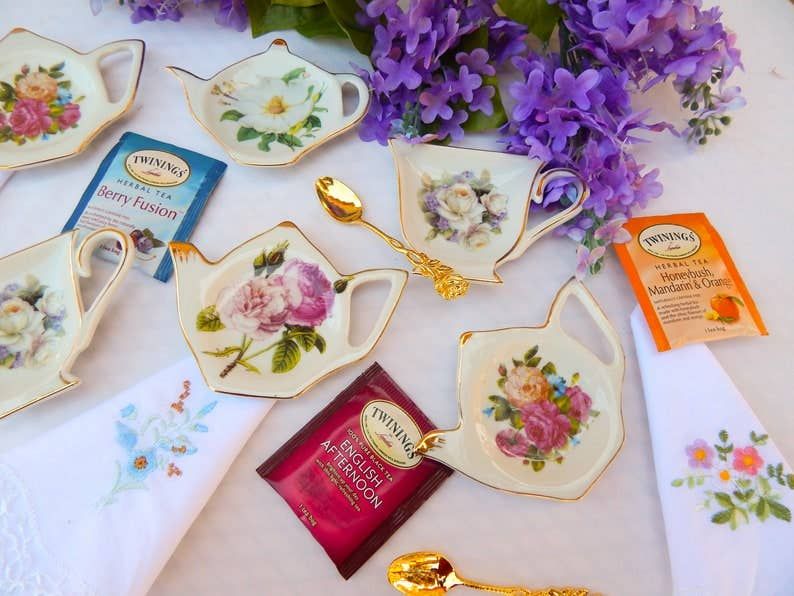 Assorted tea sets, tea bags, spoons, and floral embroidered handkerchiefs, with purple flowers on a white surface.