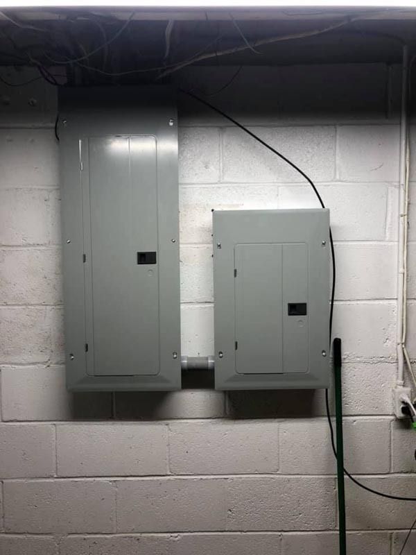 Two gray electrical panels mounted on a white cinder block wall in a basement.
